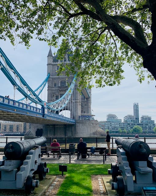 Tower bridge London with a tree and people sitting on a bench near the river looking out at the bridge.