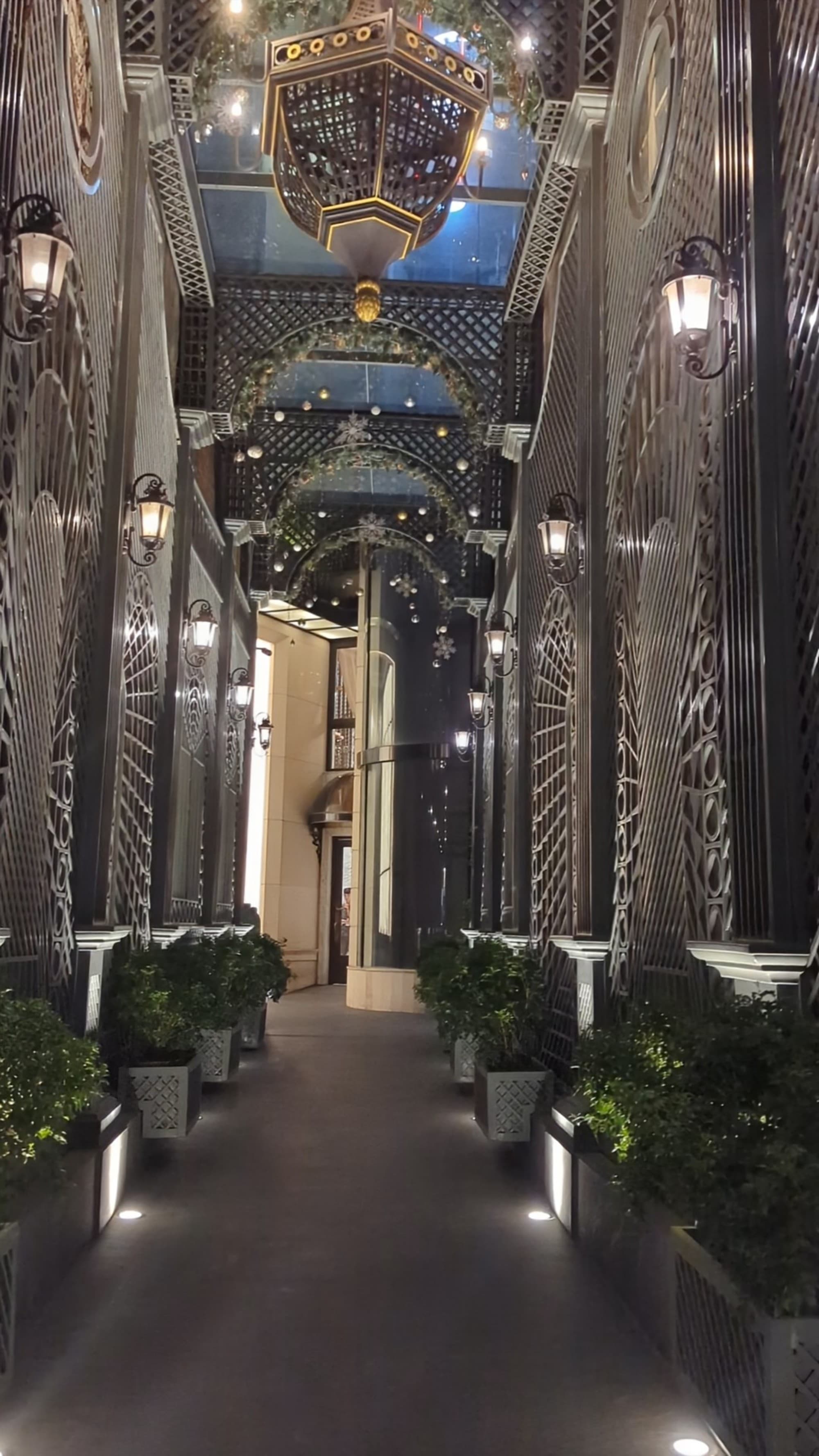 A view of a hotel entrance hallway complete with accent lighting, rectangular planters with green bushes, elaborate chandeliers lined in a row and decorative metal detailing on the walls.