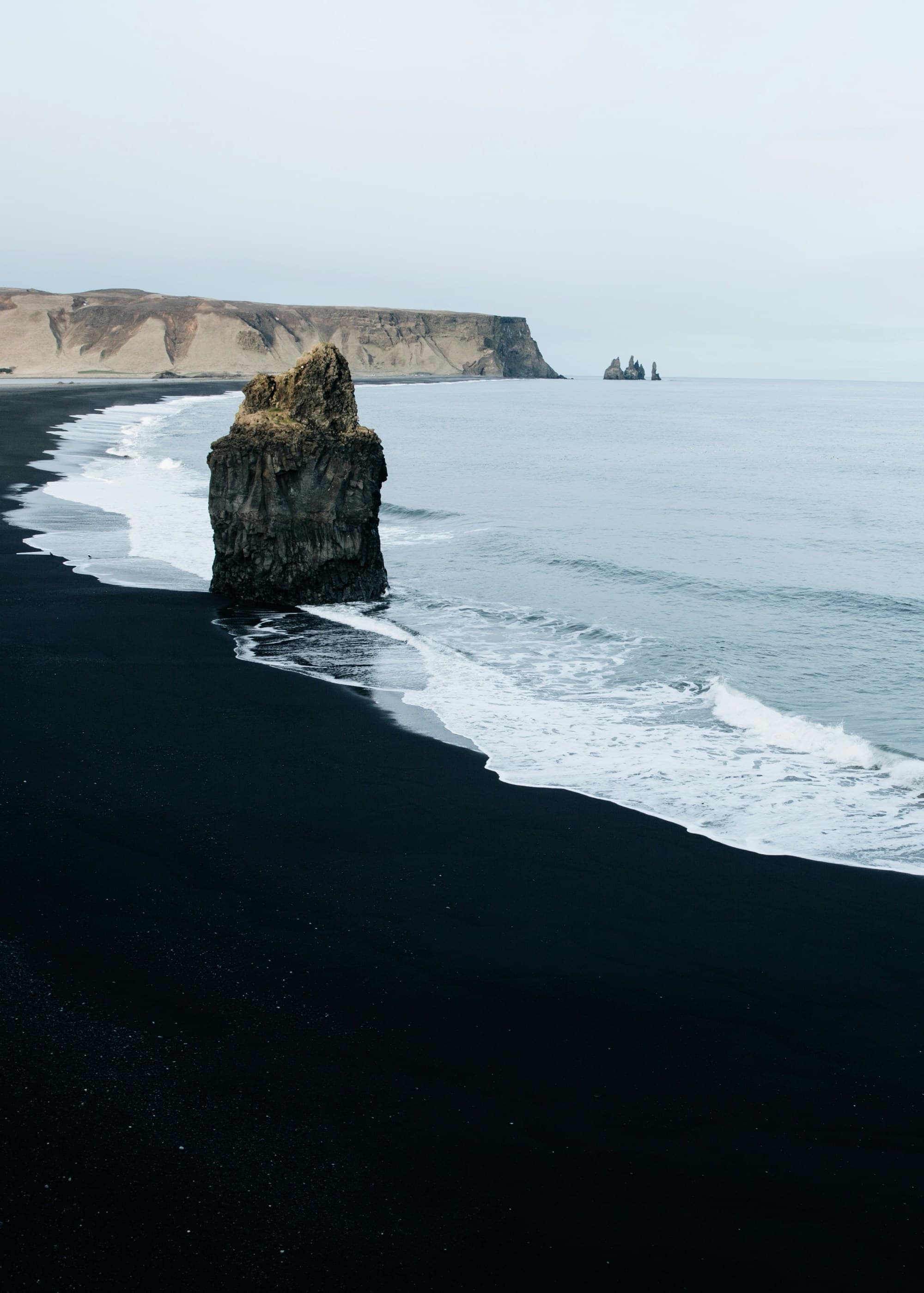 A picture of a black sand beach with waves rolling in on the shoreline. There is a large boulder in the middle of the scenery, and a rocky cliff in the background.