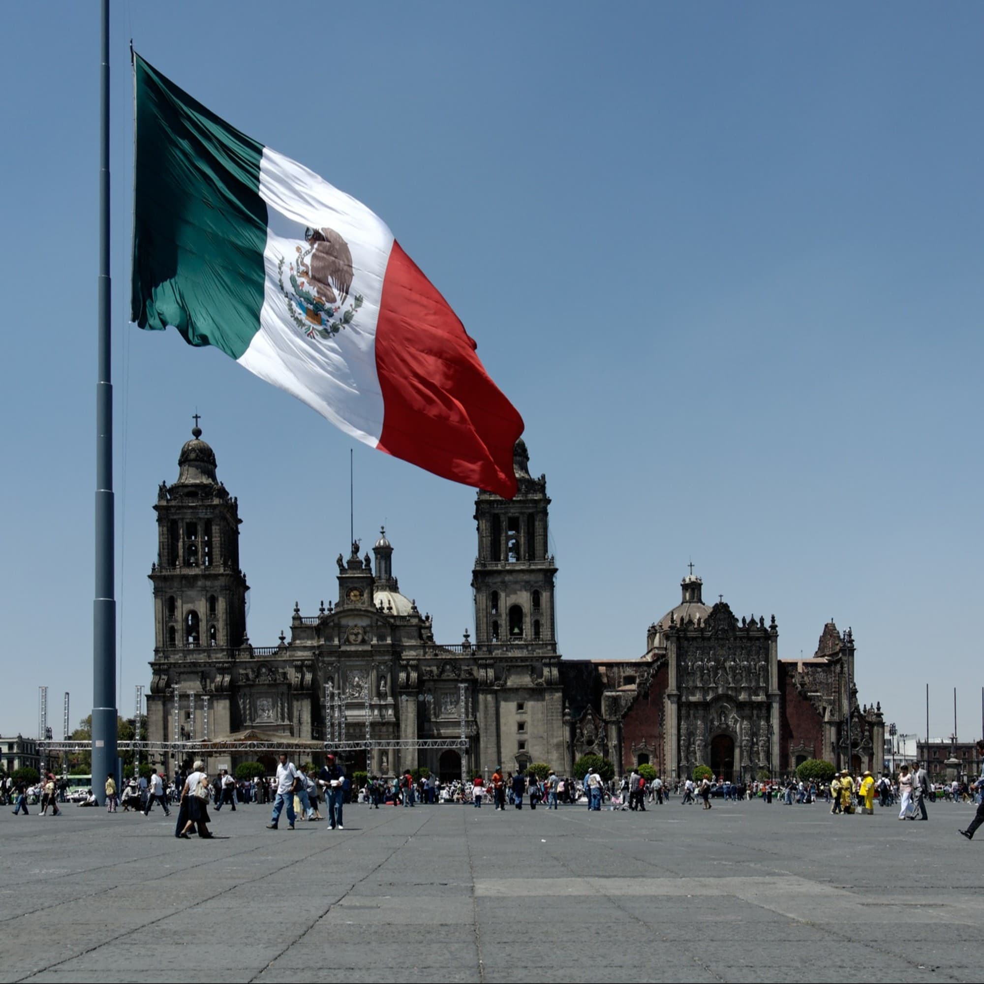 A view of the flag in Mexico in front of people walking near a grand stone building.