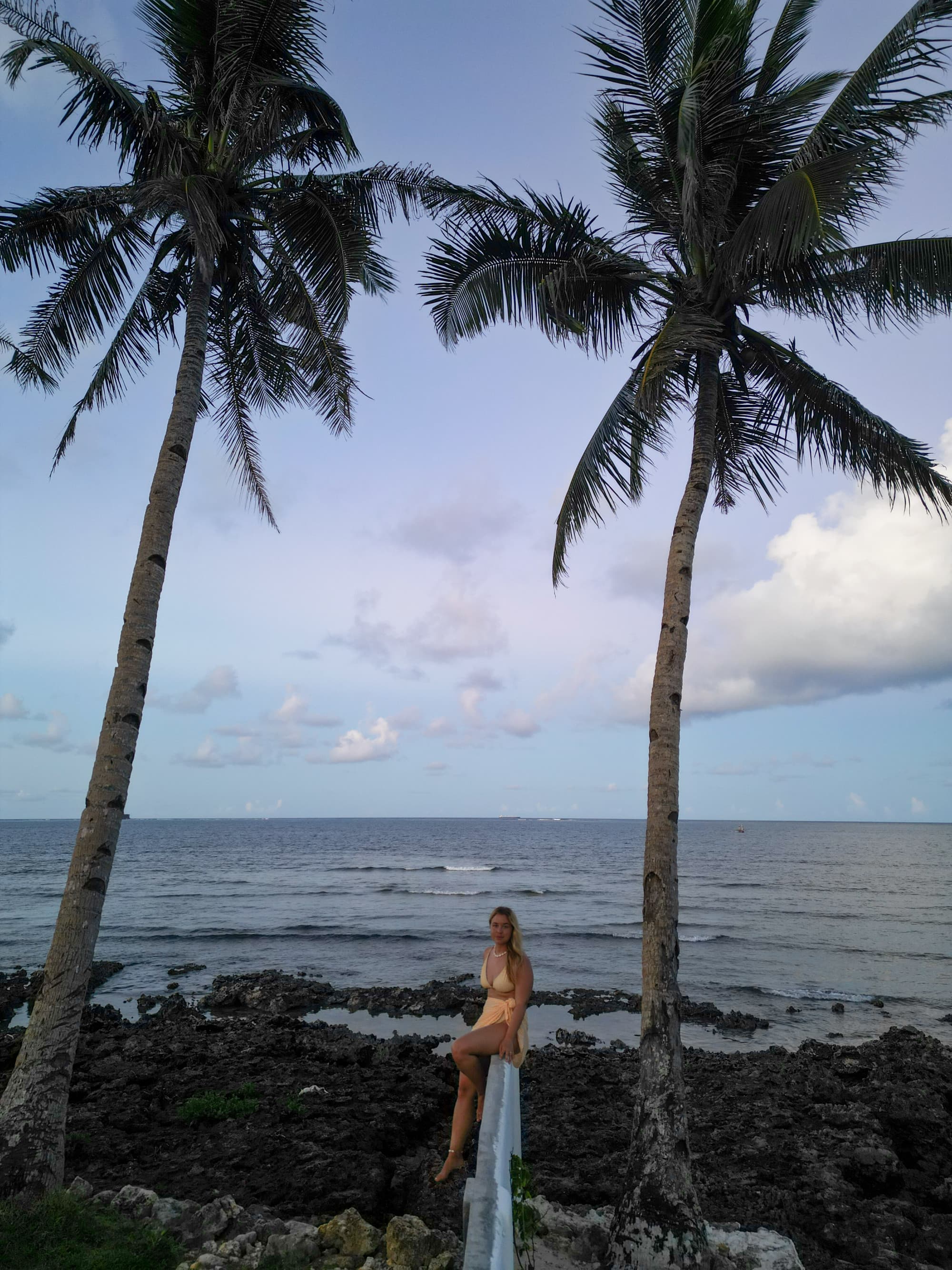 A girl posing near the beach during sunset under two tall palm trees.