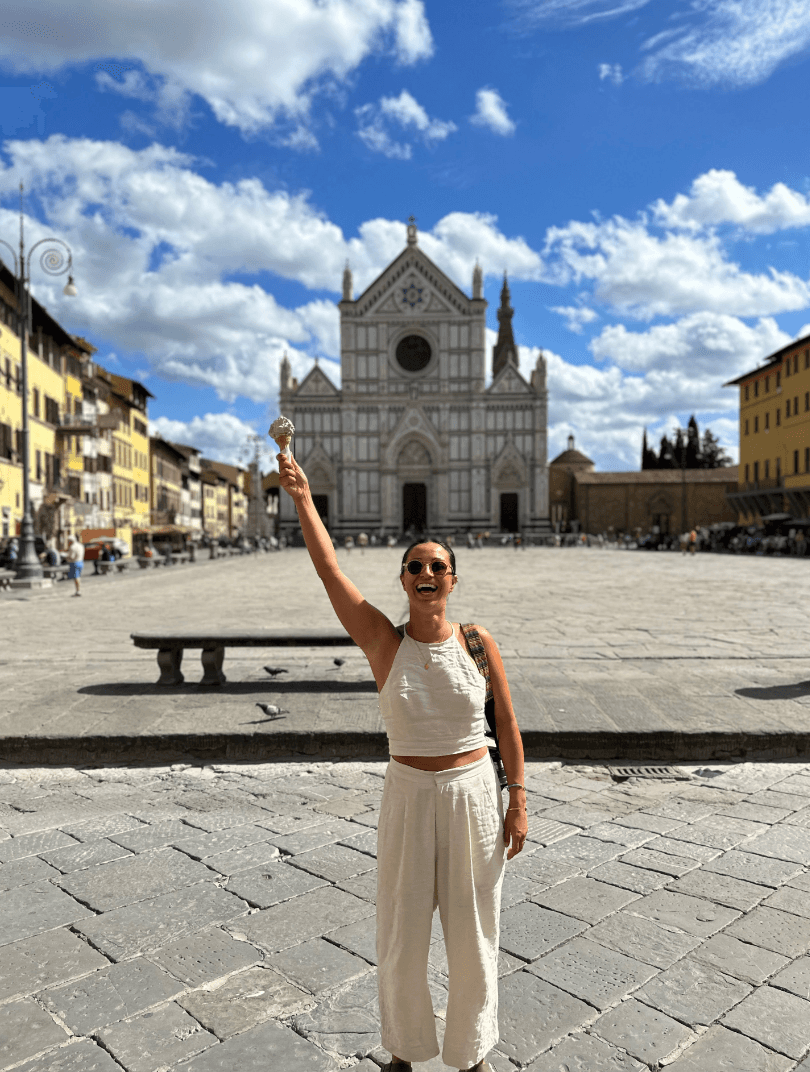 An image of Janet wearing a white outfit and holding a gelato up in the air in front of a building in Florence, Italy.