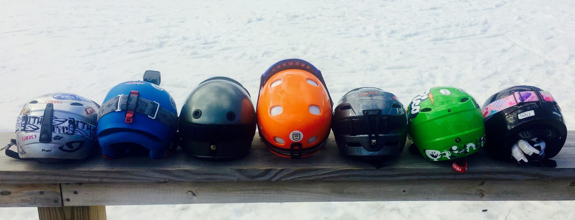 A picture of seven different colored ski helmets lined up on a wooden bench.