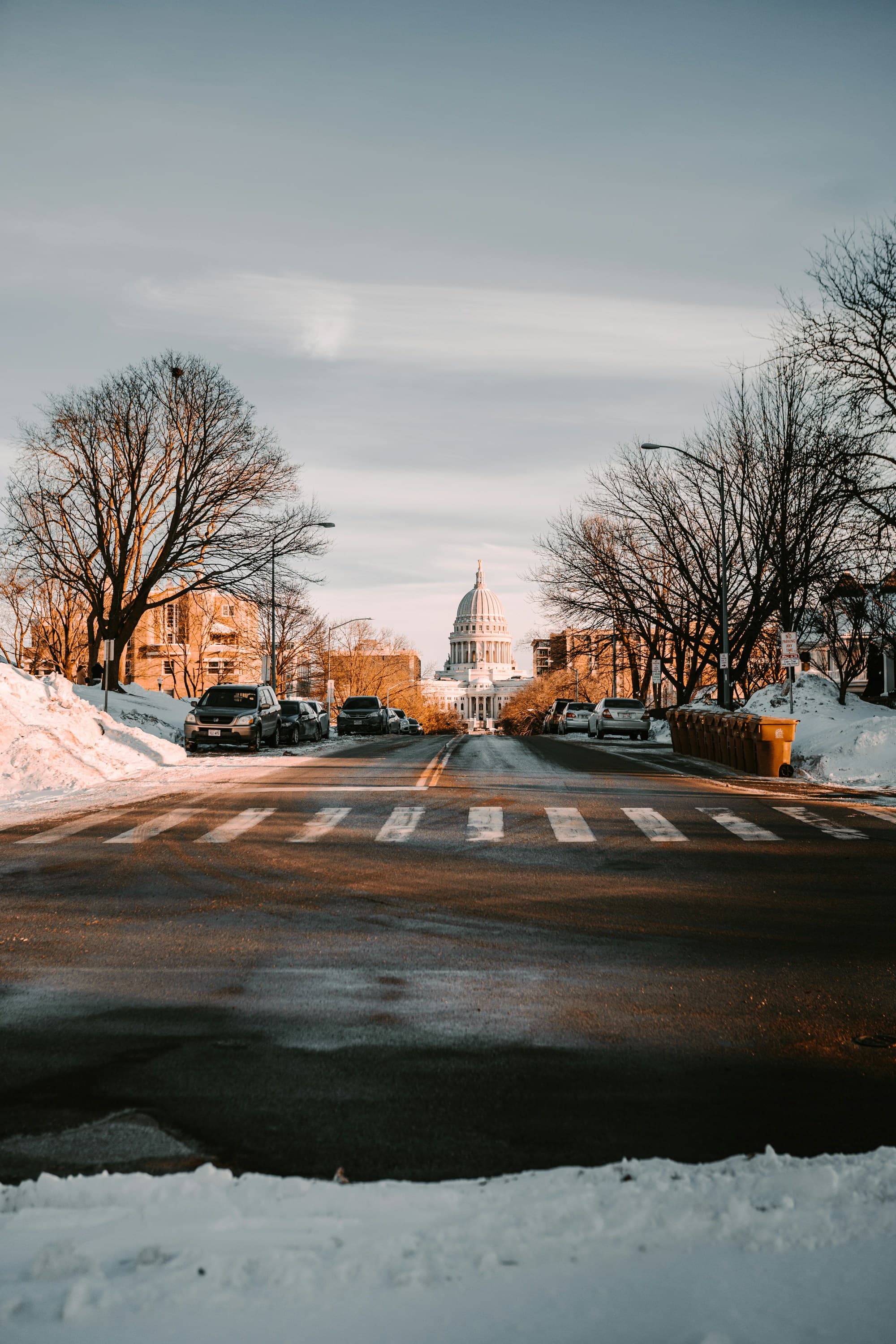 Cars parked on side of the road with bare trees and snow.