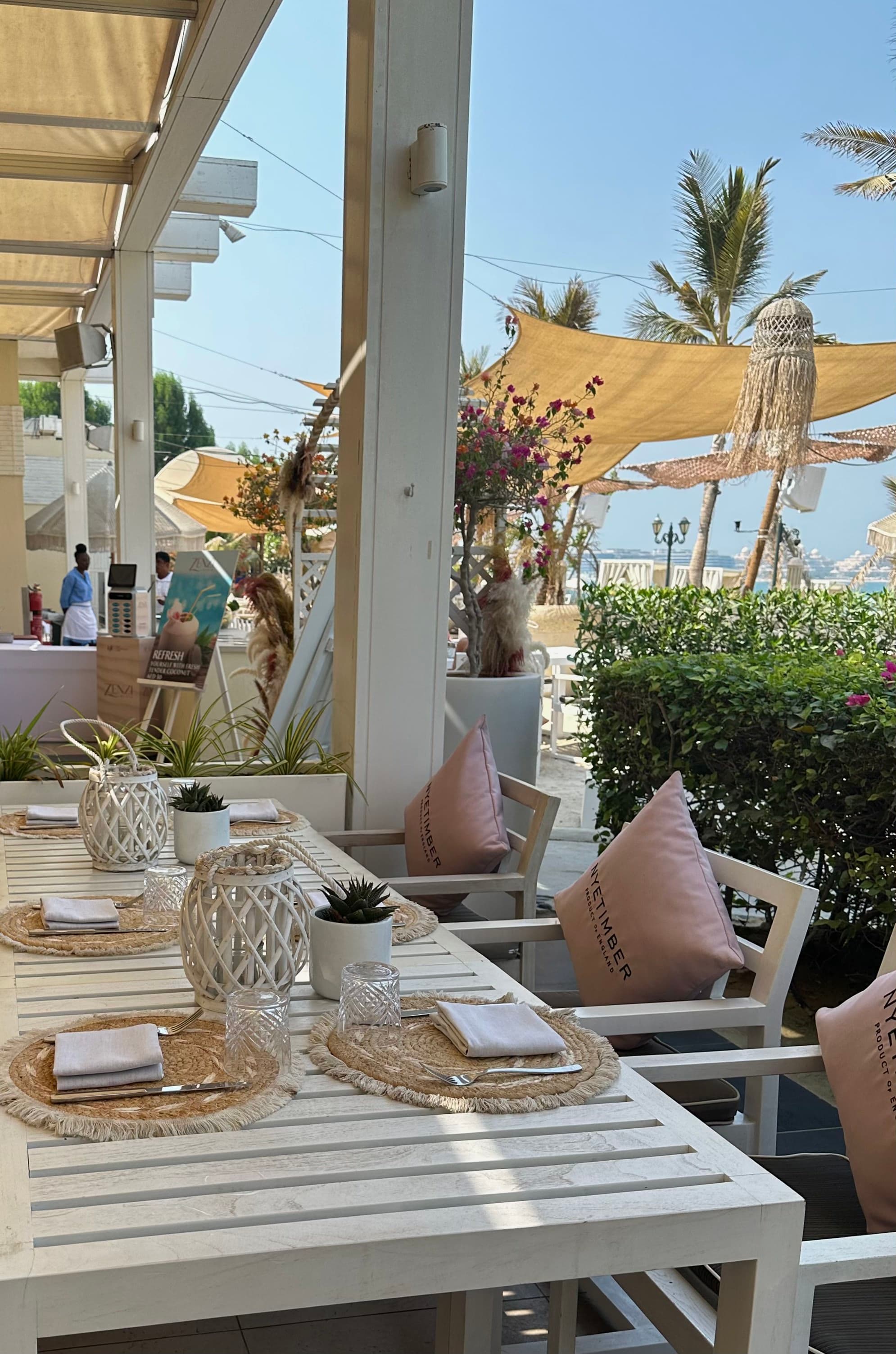 A view of a beautiful outdoor cafe with white tables and chairs that have pink pillows, woven table clothes and white lanterns. There are also sun coverings and palm trees in the background.