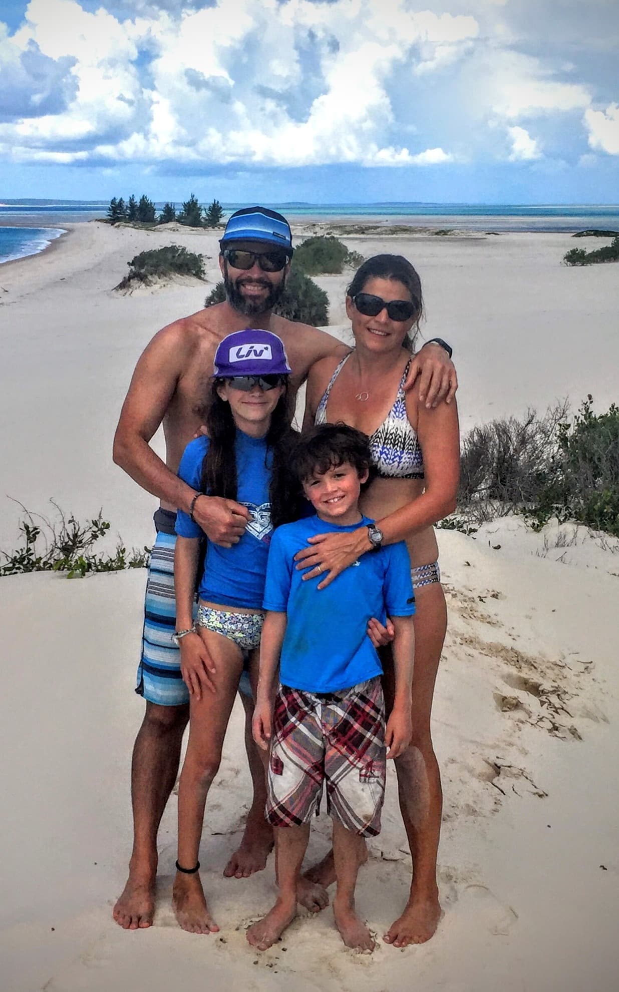 A family of four wearing swim wear posing for a photo on the beach with sand and brush in the background.