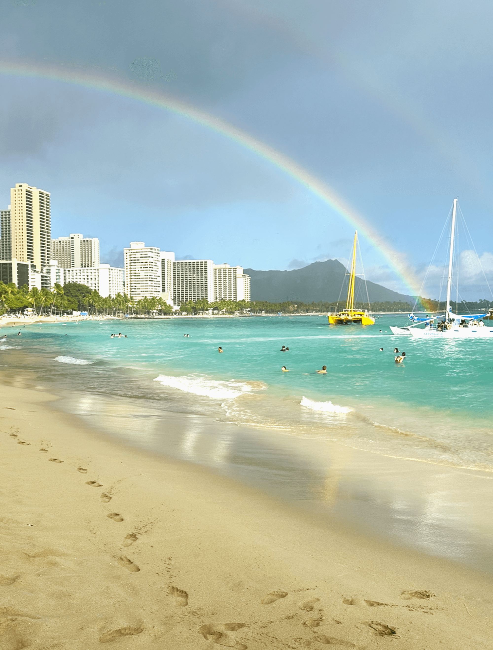 The image depicts a colorful rainbow arching over a beach with swimmers and boats, set against a cityscape and mountains.