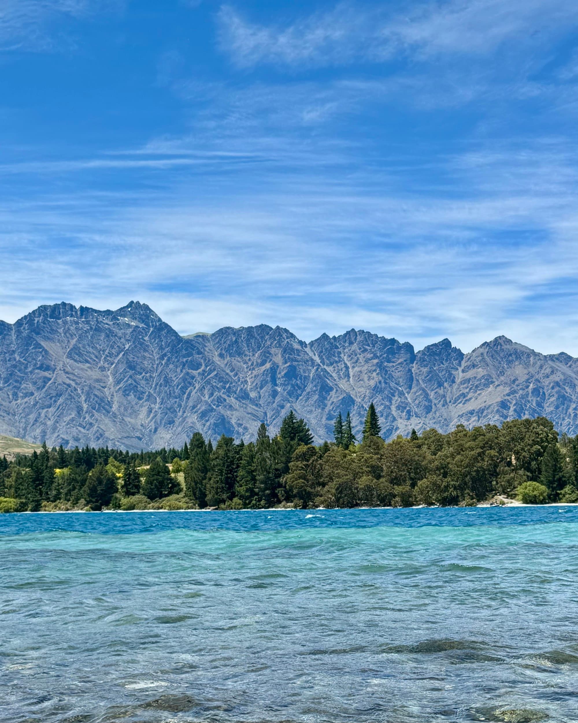 A vibrant landscape with a clear blue lake in the foreground and rugged mountains in the background under a partly cloudy sky.