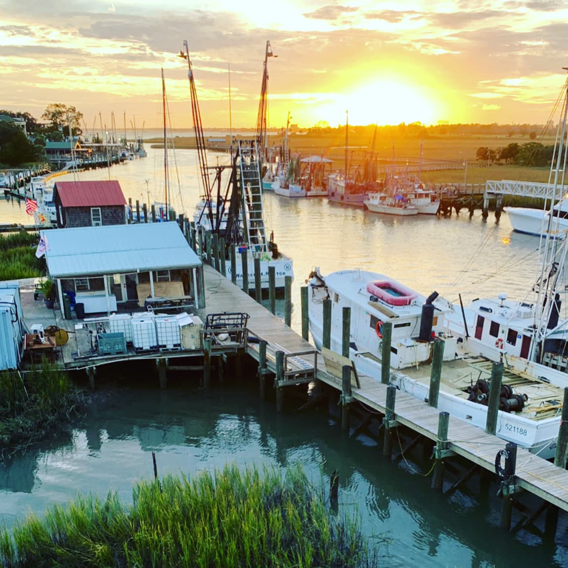 A Charleston sea port along the docks is dotted with boats as the sun sets behind the horizon at dusk.