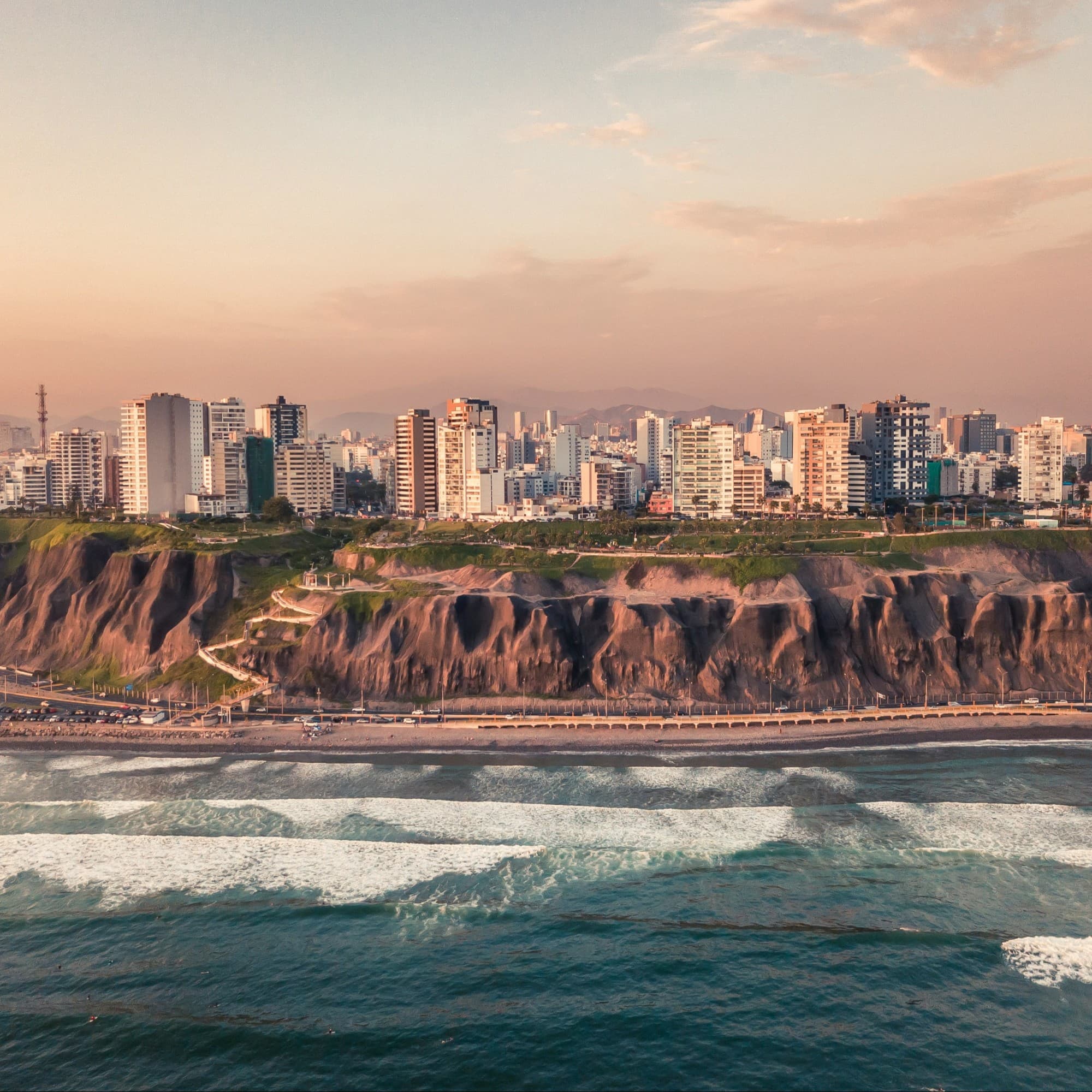 A coastal cityscape with buildings along the shore, cliffs, and a beachfront with waves breaking in the foreground.