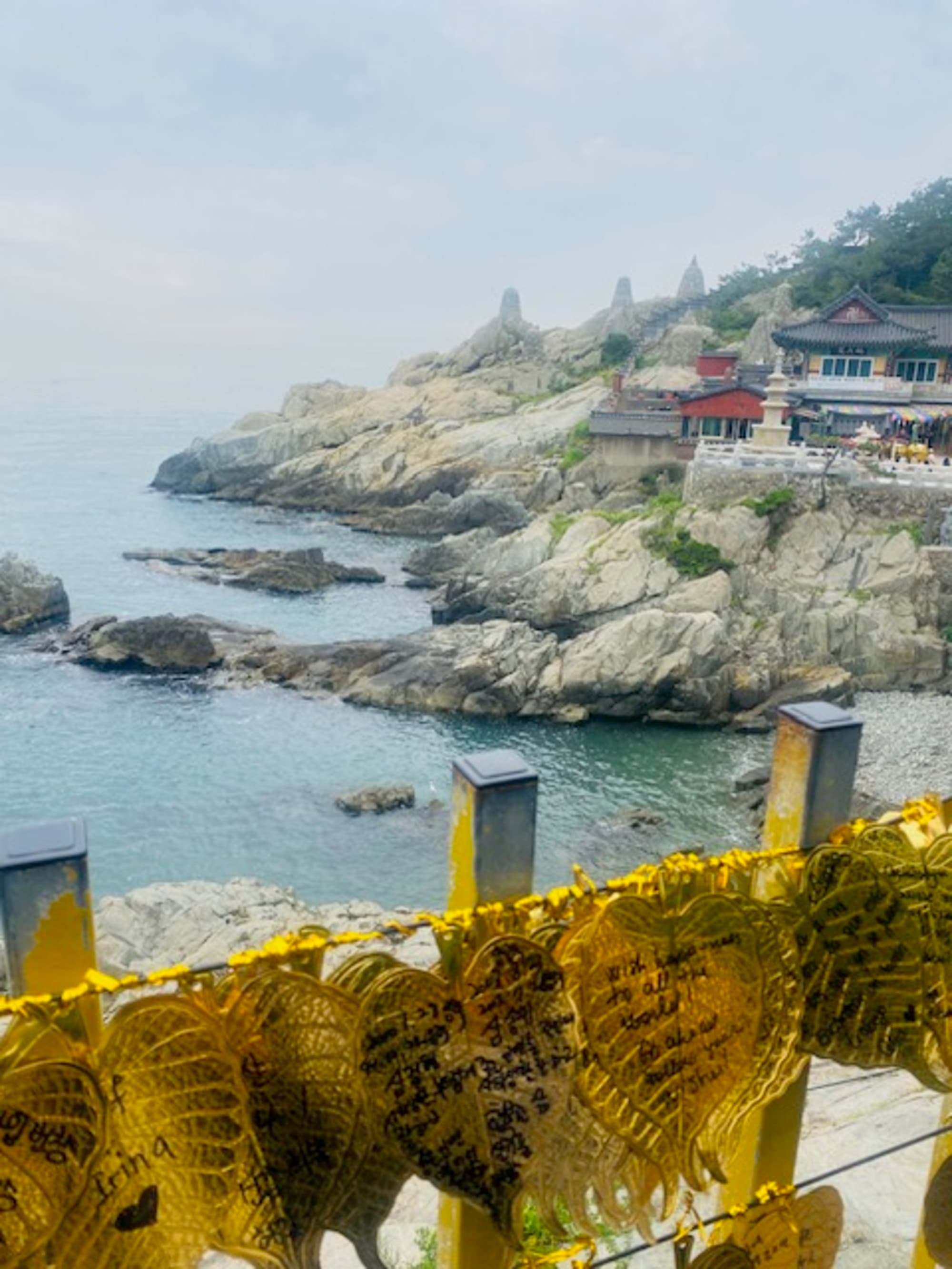 The image captures a rocky coastline with a structure and yellow ribbons in the foreground against hazy skies.