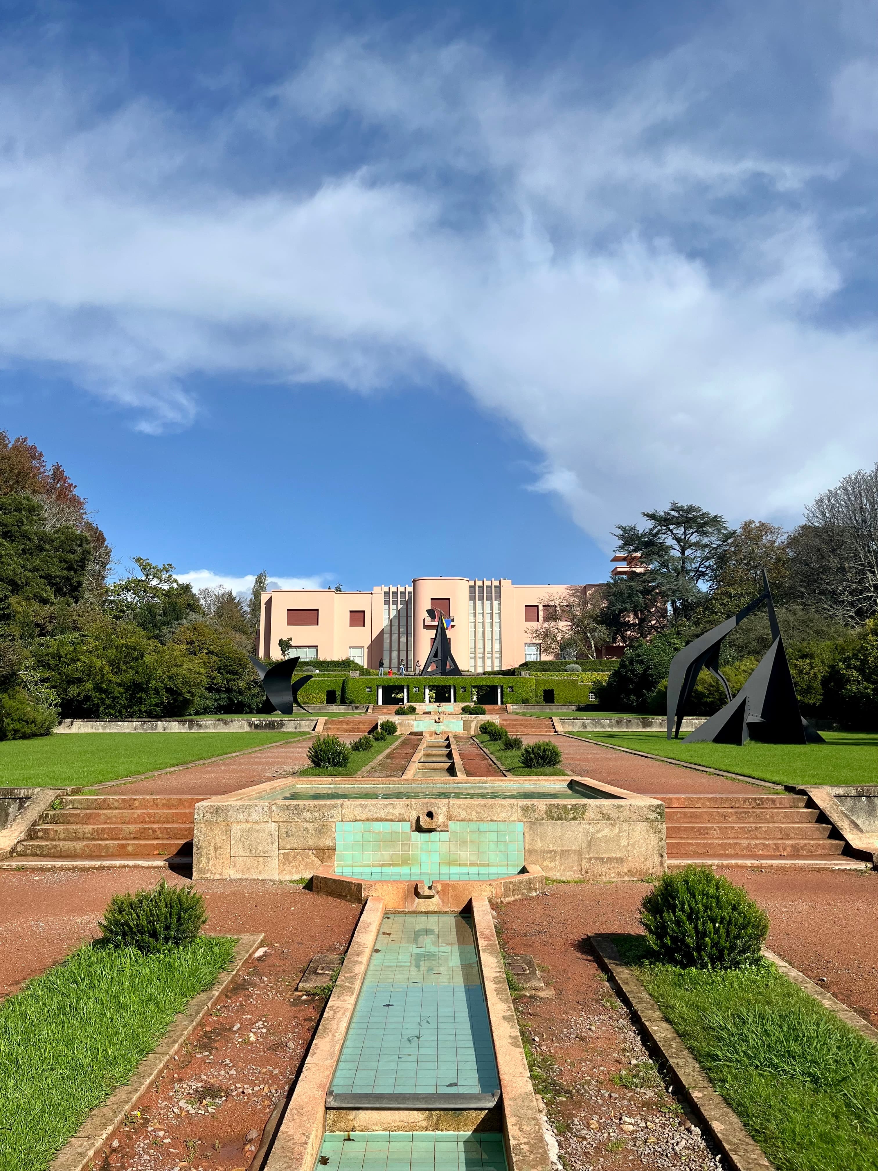 The image portrays a symmetrically designed garden with a water feature leading to a building adorned with sculptures under a clear blue sky.