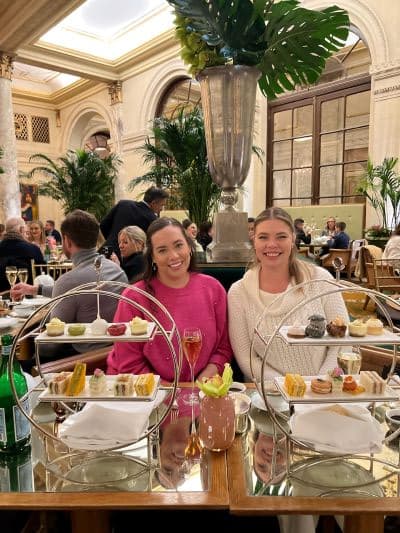 Two women at a hi-tea in a brightly lit restuarant with green palm leaf arrangements and a large skylight.