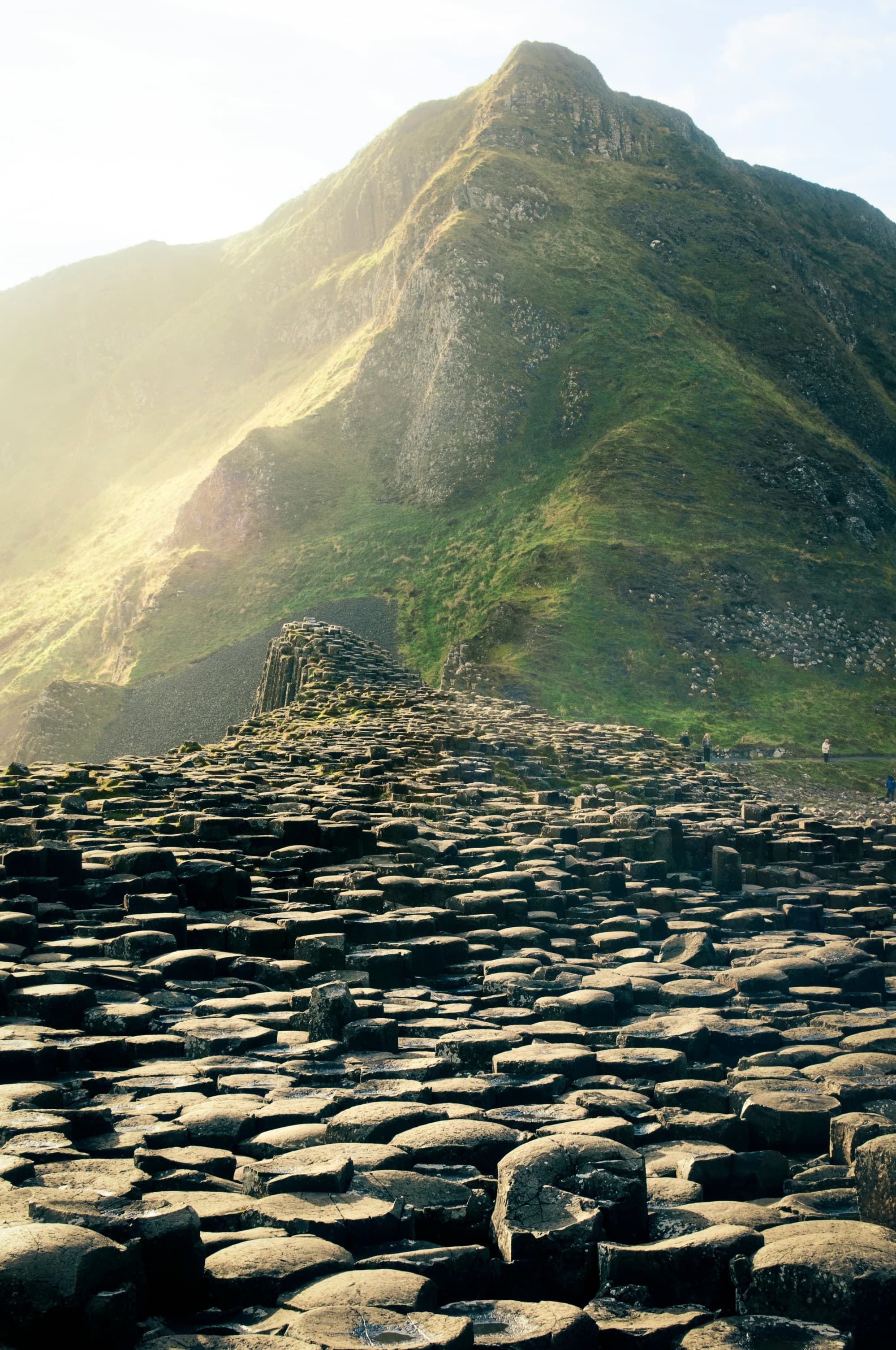 The image showcases a natural landscape with unique hexagonal rock formations and a sunlit green mountain in the background in one of Ireland's National Parks.