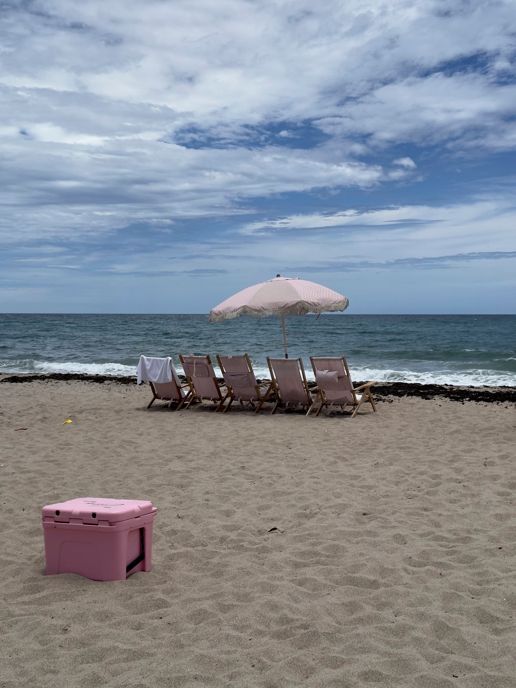 The image portrays a tranquil beach setting with a pink cooler, four wooden lounge chairs, and a striped sand umbrella overlooking the ocean.