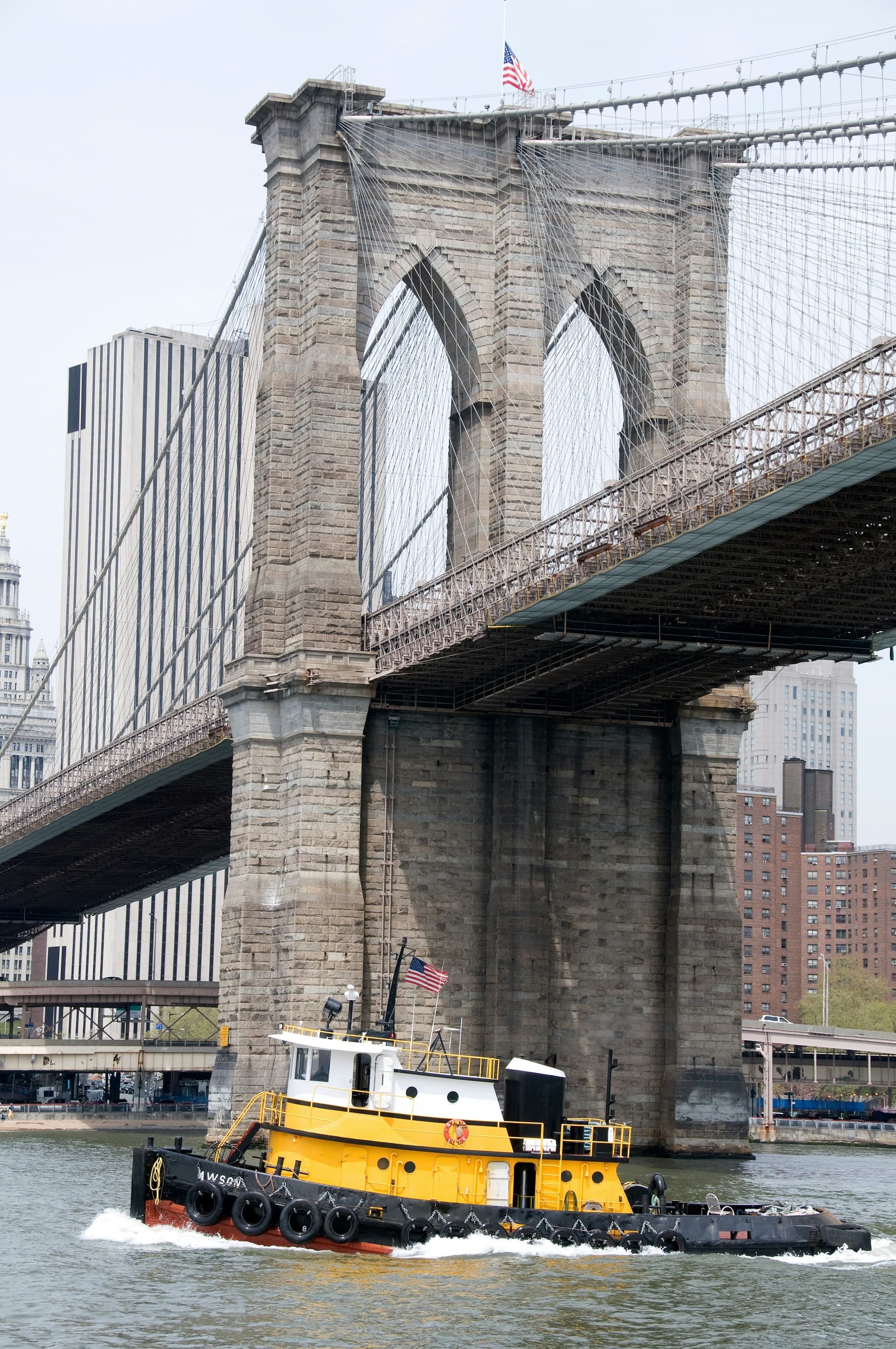 The image shows a yellow and black tugboat moving on water under a large stone bridge with an American flag on top.