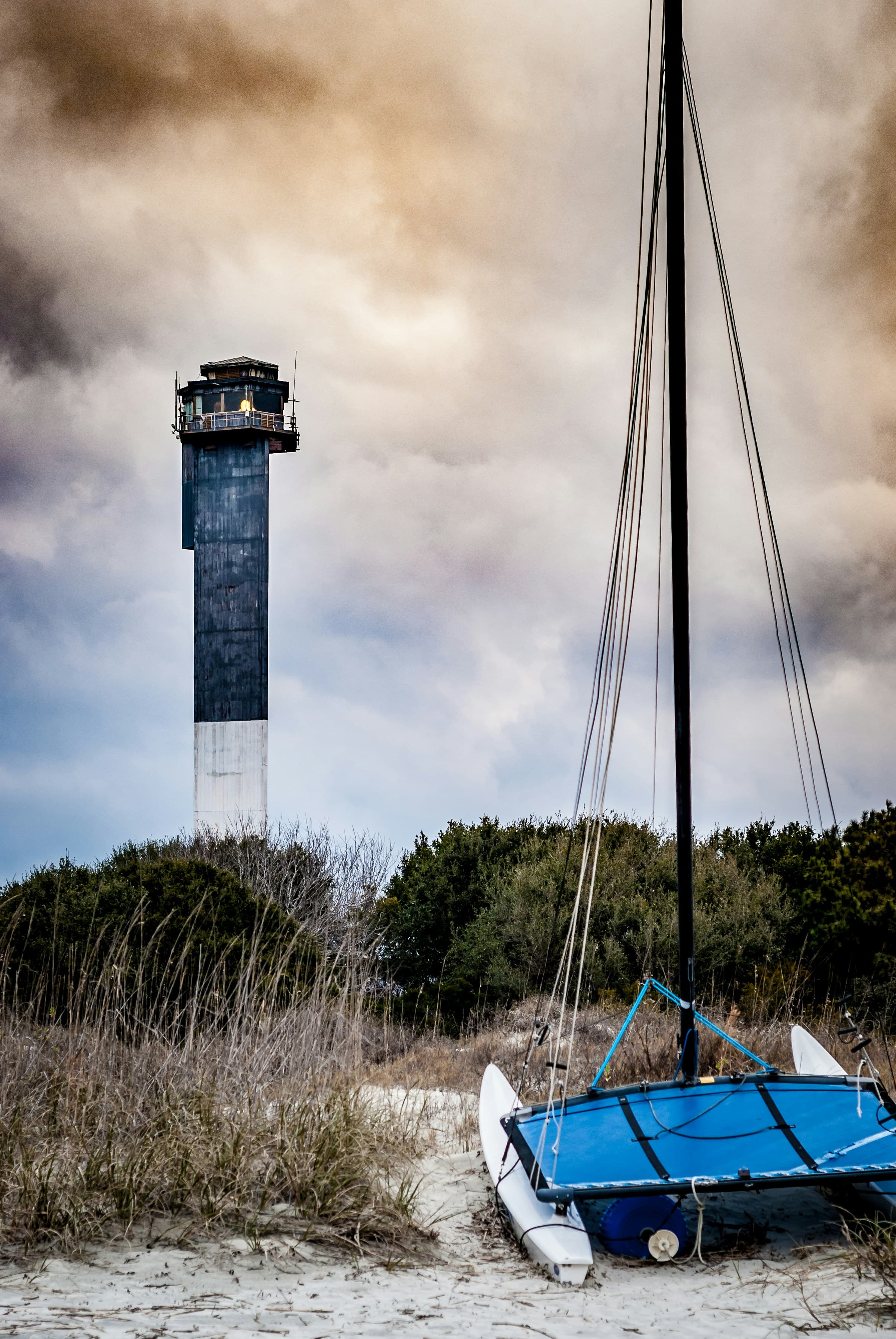 A blue boat on the sand next to a lighthouse with impressive clouds in the background on Sullivan's Island.