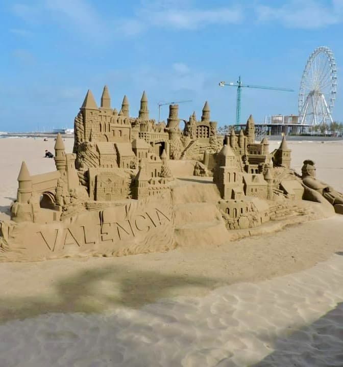 A giant sand castle on the beach with a ferris wheel and blue skies in the background.