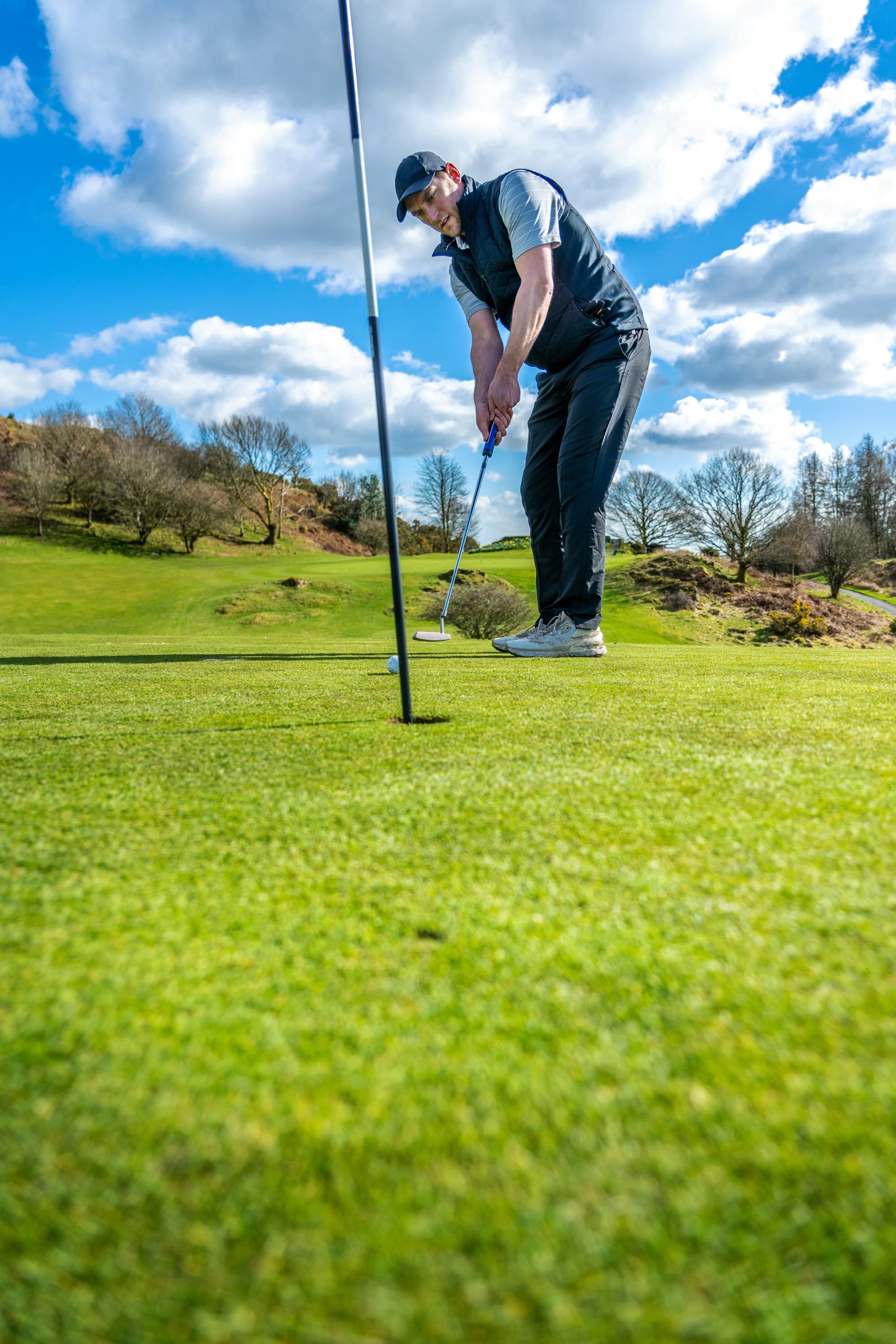 A golfer about to putt on a green, set against a hilly landscape and a partly cloudy sky.