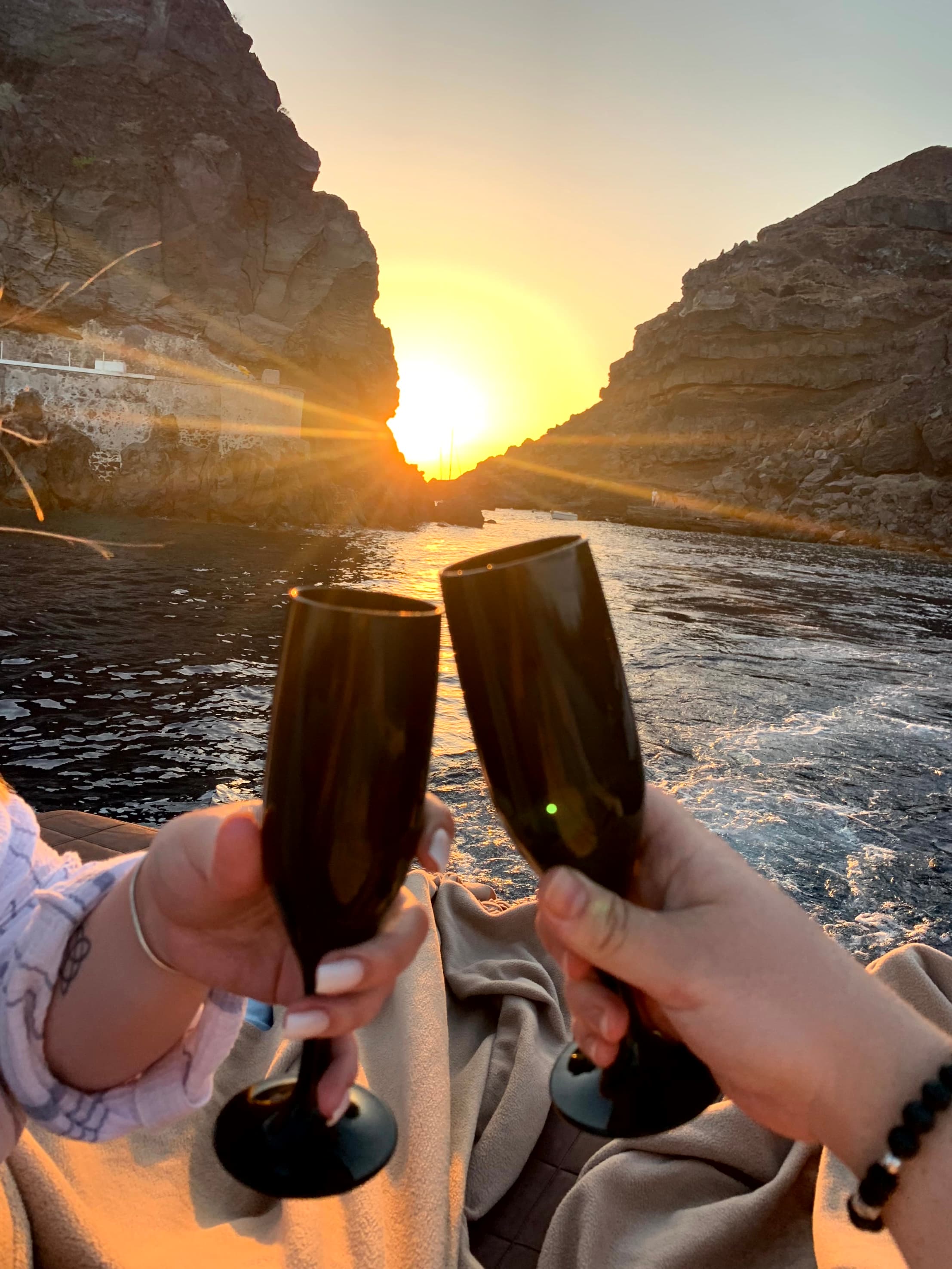 A toast of two champagne glasses in front of a blazing sunset between two craggy rock formations on the water.