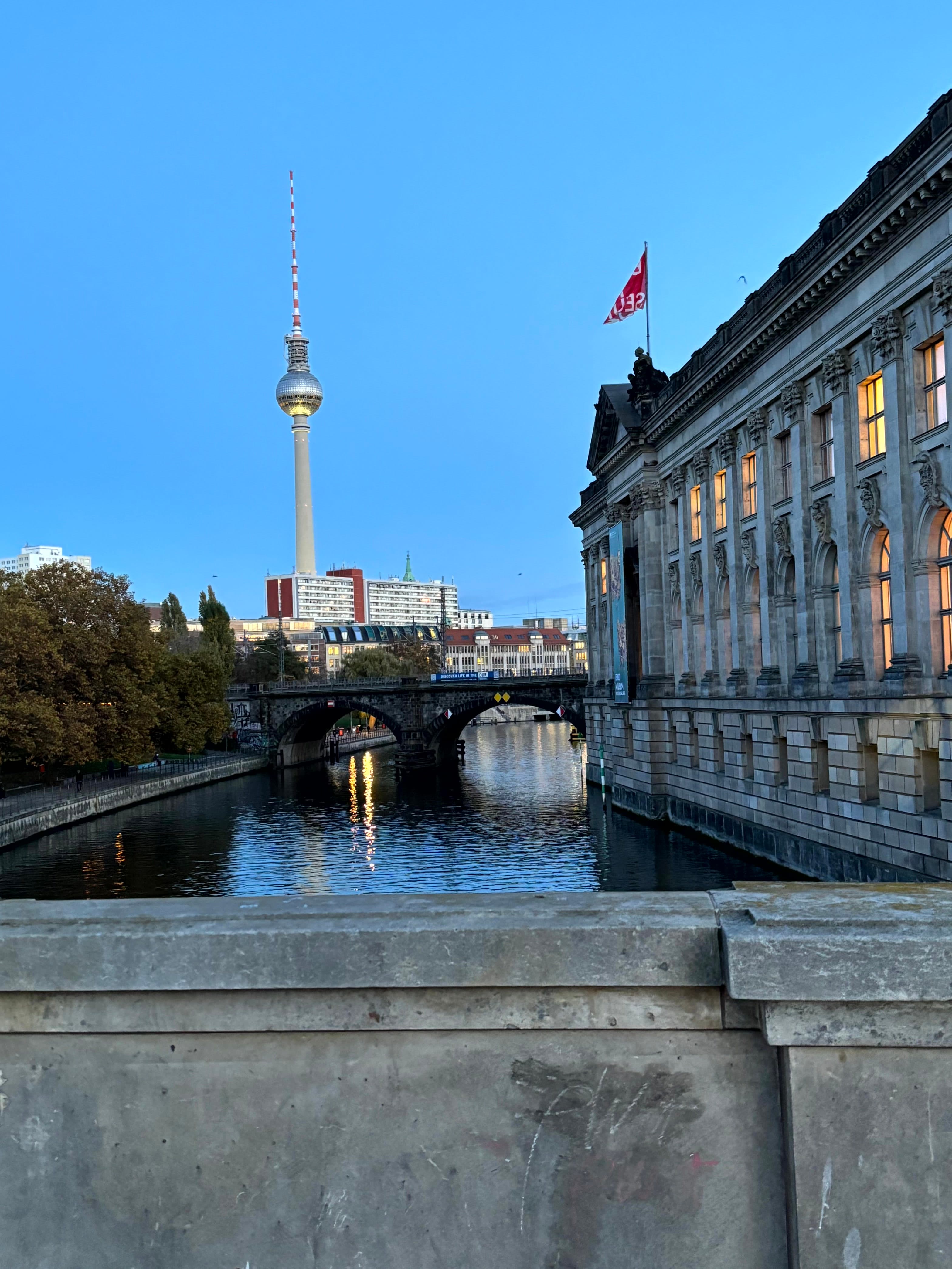 A stone building next to a river with the TV tower in the background, as seen from a concrete bridge in Berlin.