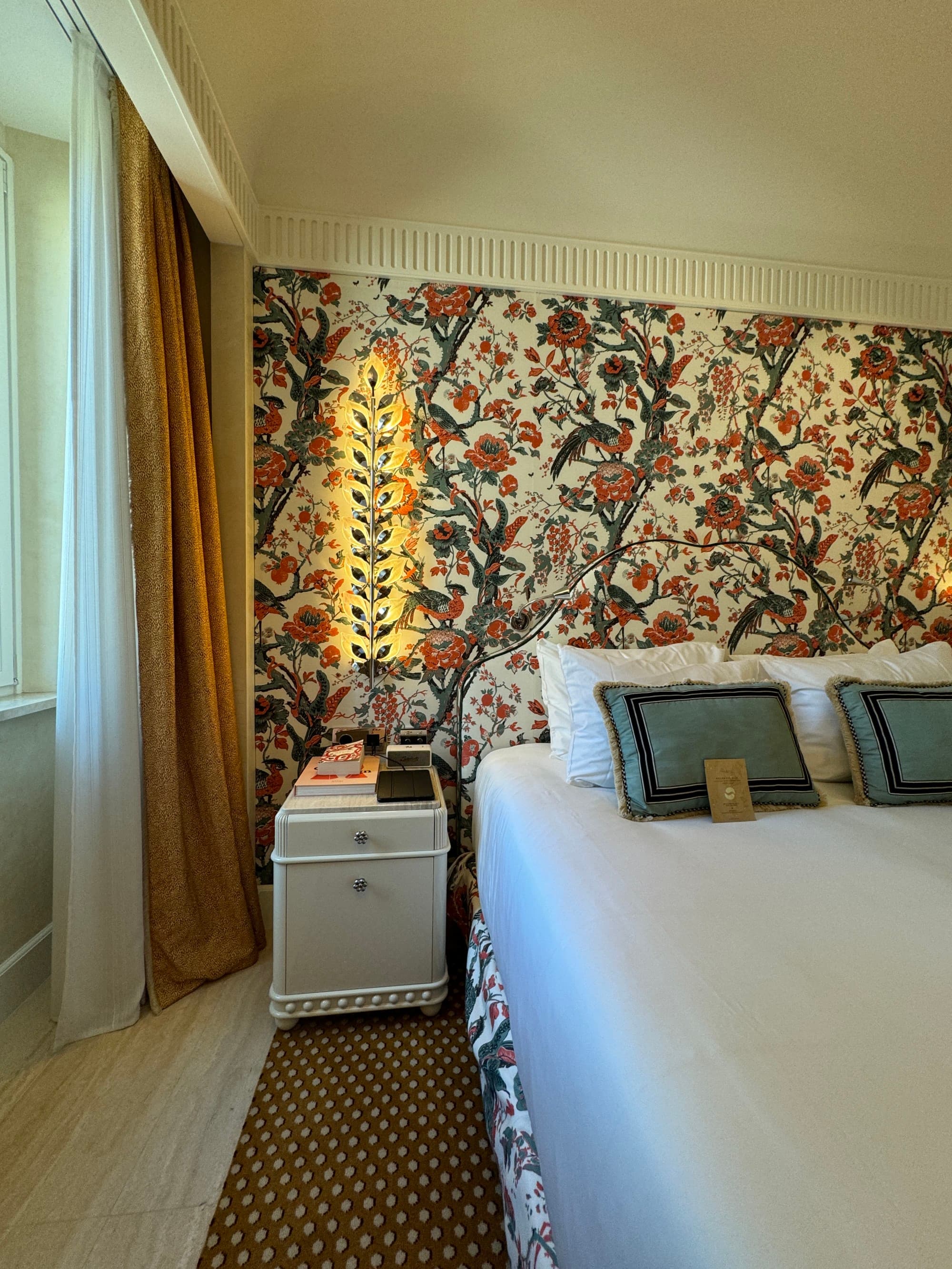 A view of the hotel room with colorful wallpaper, a white bedside table, curtains and a neatly made bed with white bedding.