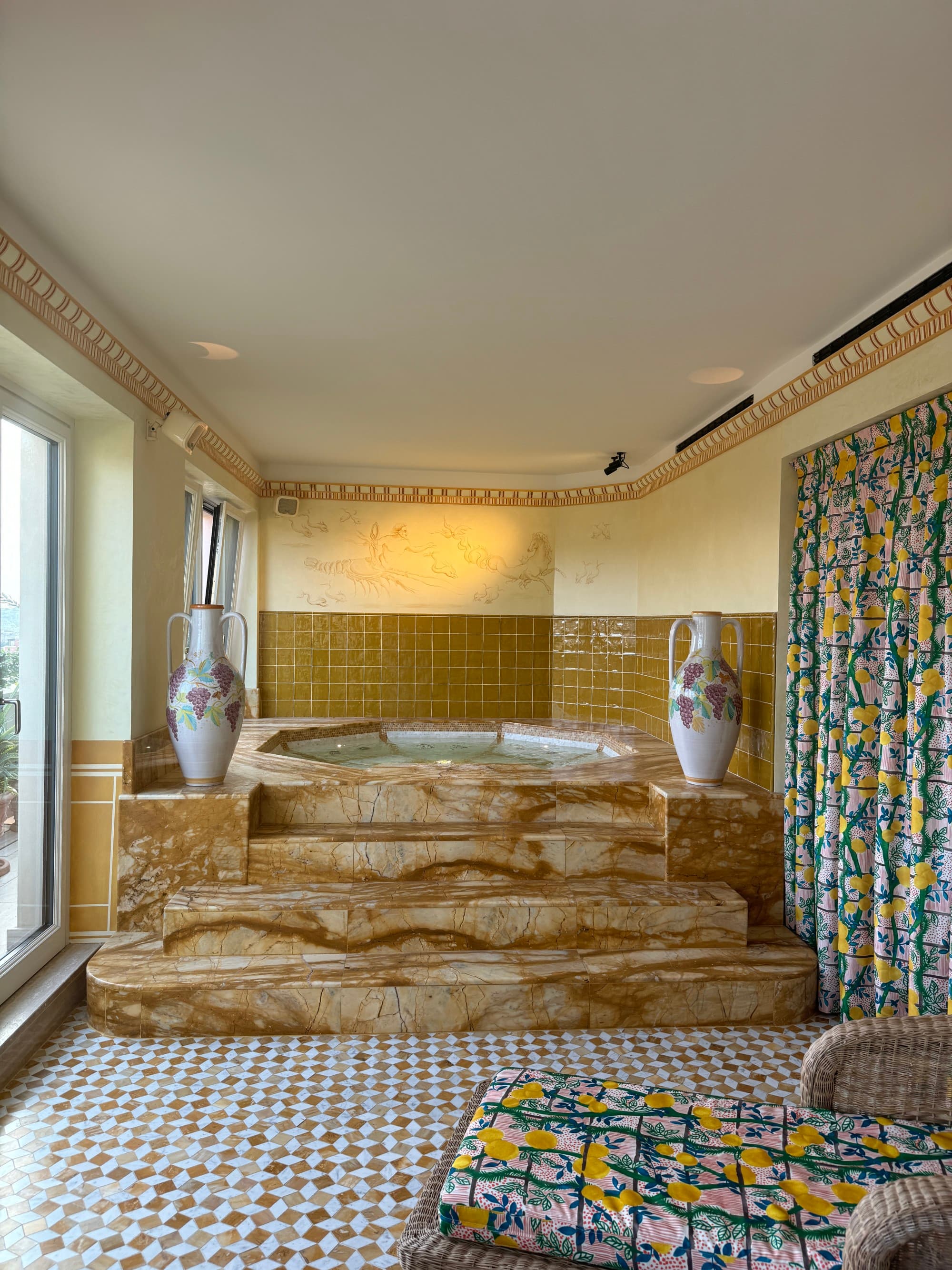 A sitting area with a chaise lounge looking into a tiled bathroom with vibrant patterned curtains, brown stone steps leading into a spa with warm lighting.