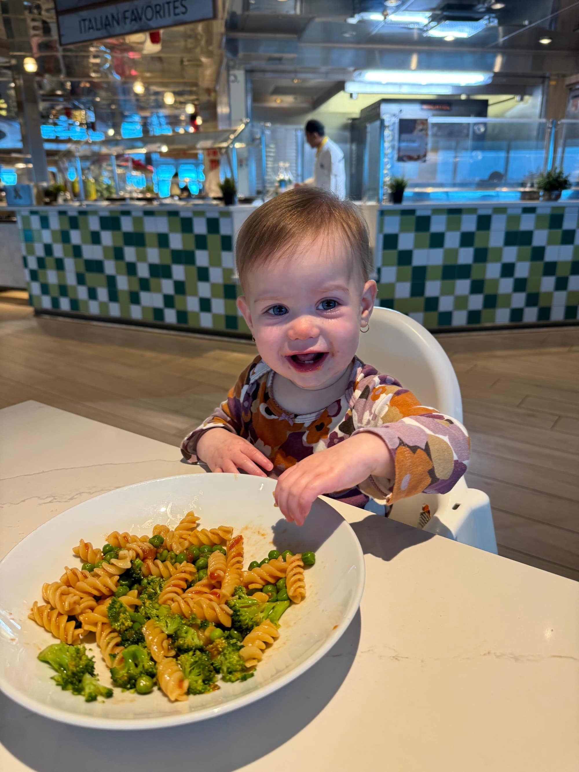 A baby sits in a high chair in a dining area with a plate of colorful pasta in front of them.