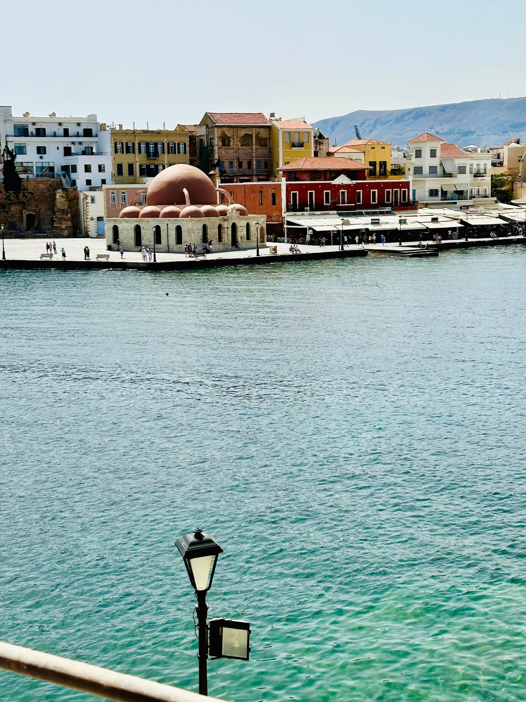 Waves gently lap the shore of a pier lined with buildings on a sun-bleached day.