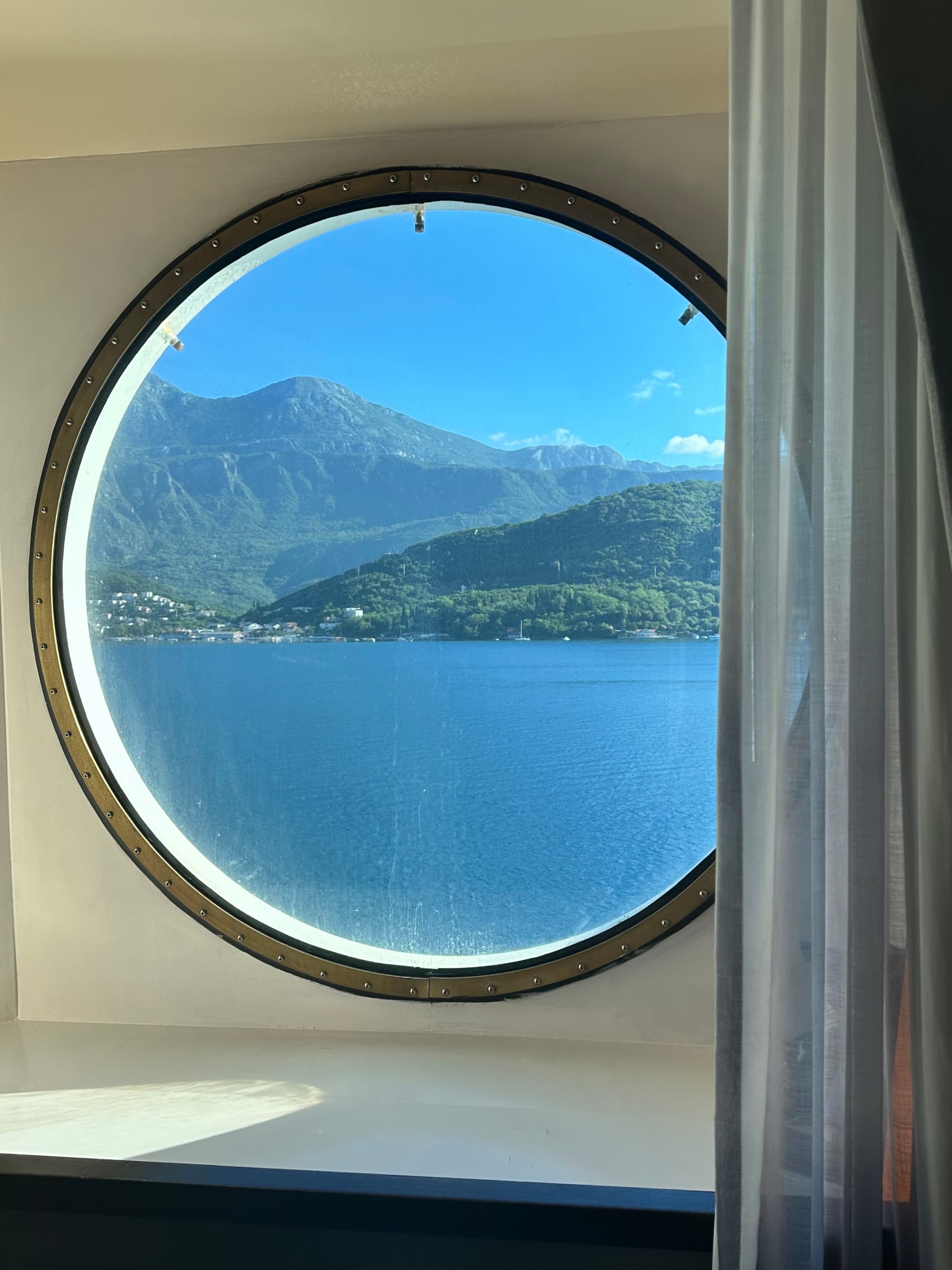 A view of the ocean shore lapping against green island hillsides through a round window aboard the ship.