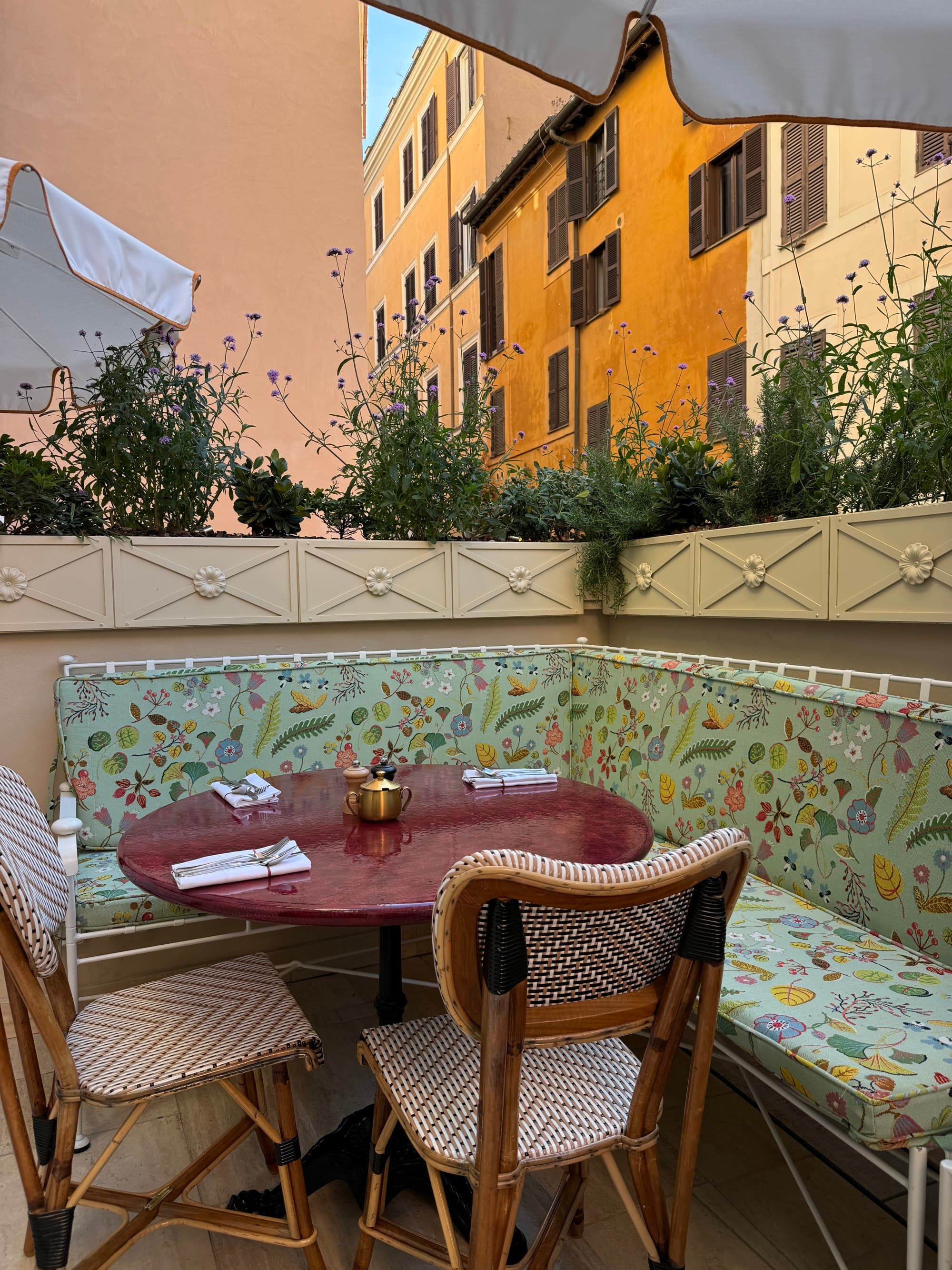 A view of a sitting area with a corner booth, a red table and two wicker chairs on a balcony with planters looking out to the city.