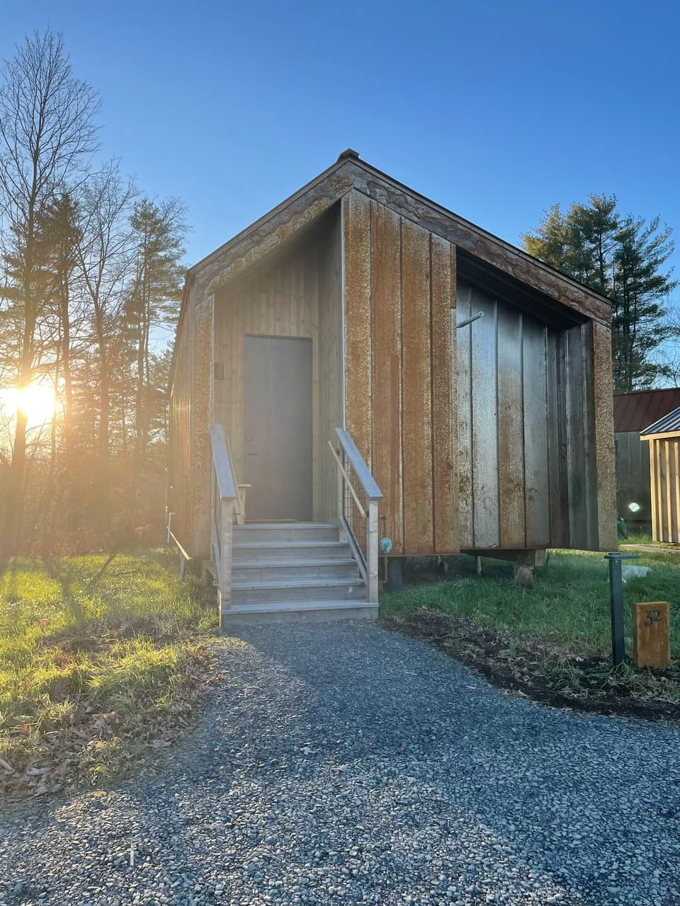 BOWERCABIN, a wooden cabin with grass and trees around it with the sun rising behind it against a blue sky.