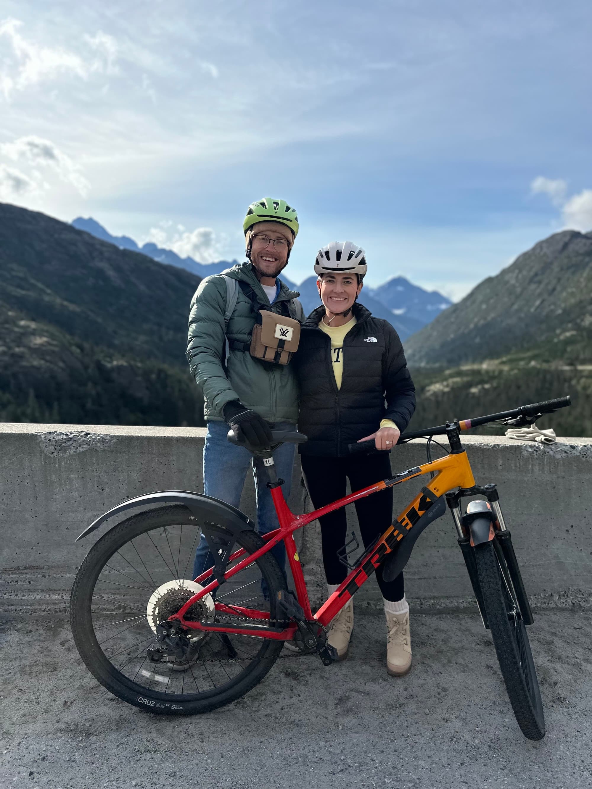 Two people posing for a photo with a bicycle with mountains in the background