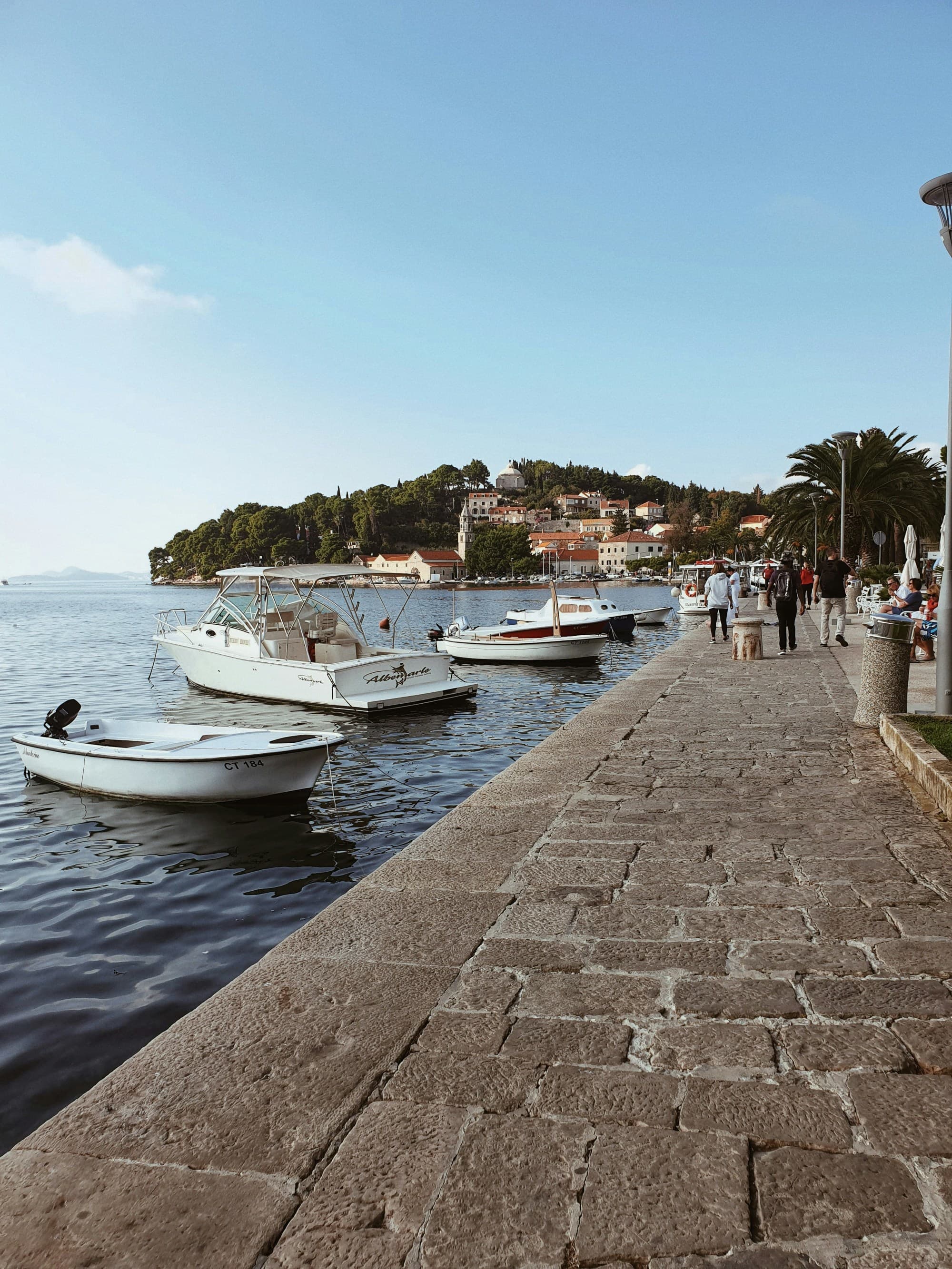 A picturesque promenade by the water with moored boats and a hillside town in the background.