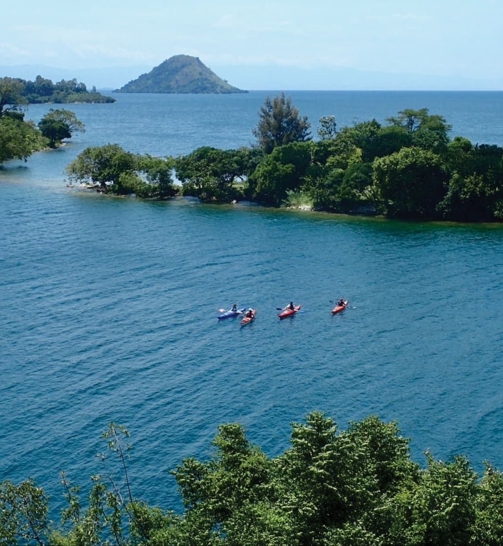 Kayakers on the beautiful Lake Kivu, a large blue water body with strips of trees jutting into it and a mountain in the distance.