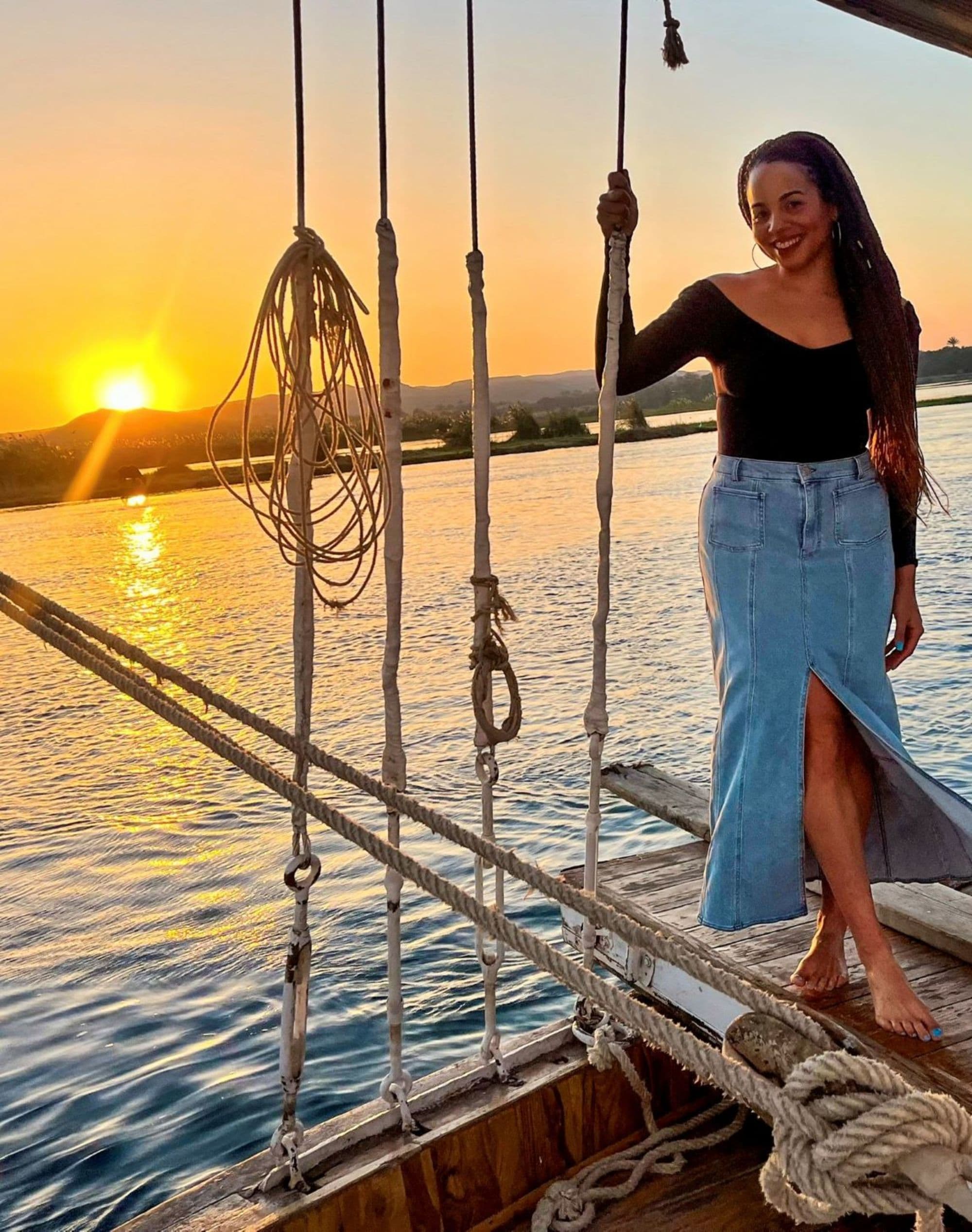 Elizabeth in a black top and denim skirt on a boat’s deck at sunset, surrounded by ropes and rigging.