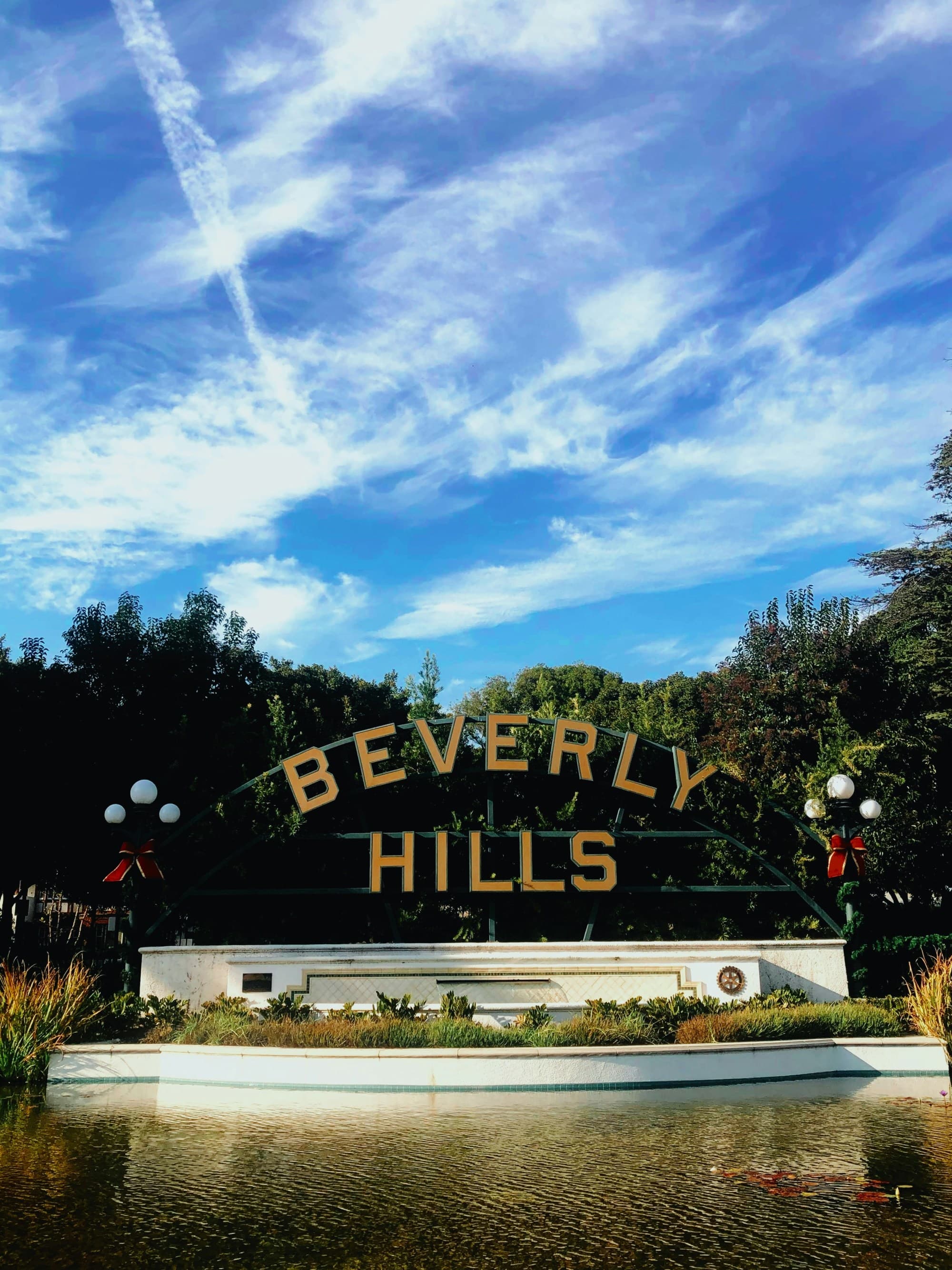 The image depicts the famous Beverly Hills sign amidst lush greenery under a clear blue sky, leading to hotels near Rodeo Drive.