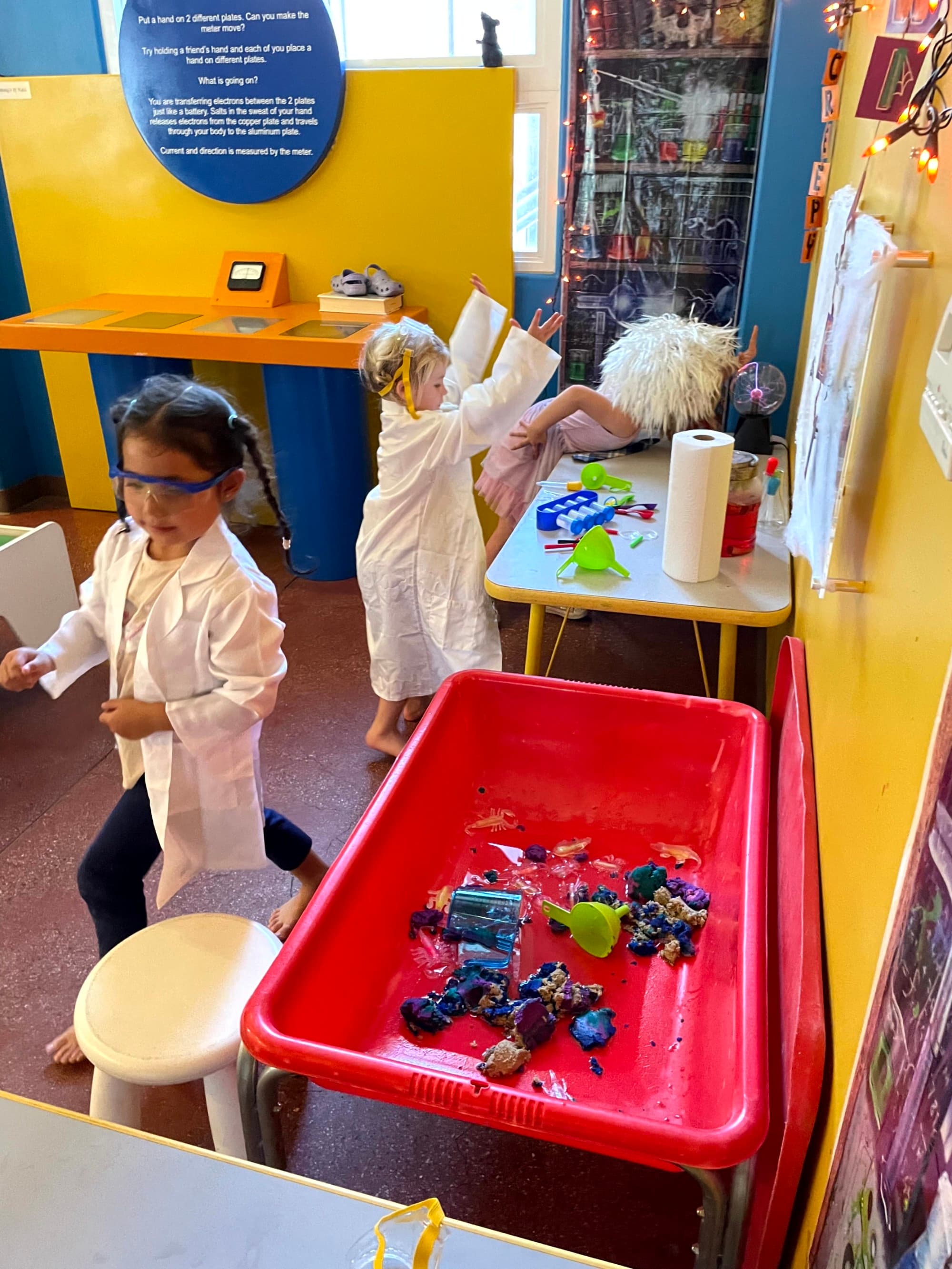 Two children in white lab coats enjoying an activity at a museum with various art supplies on a table.