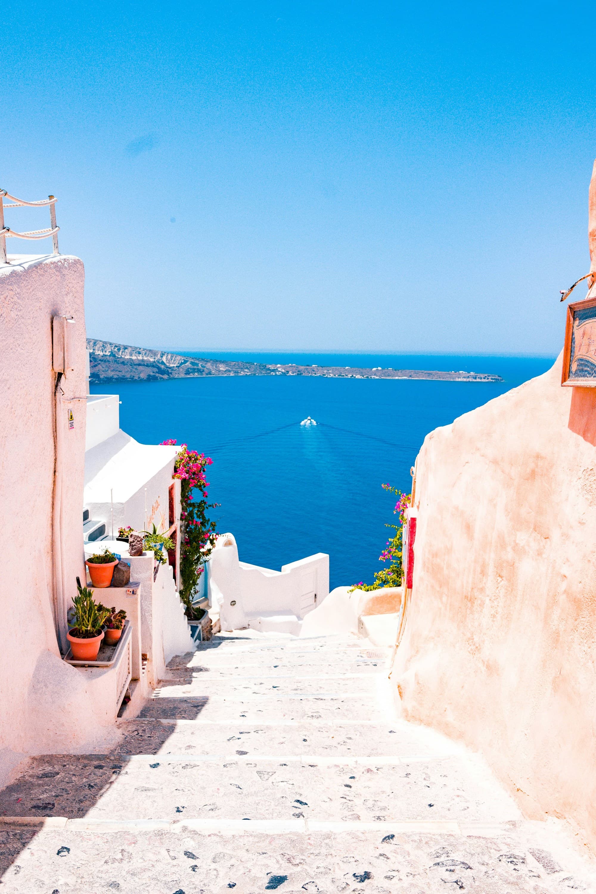 A white stone staircase between pink and white Greek buildings leading down to a brilliant blue ocean.