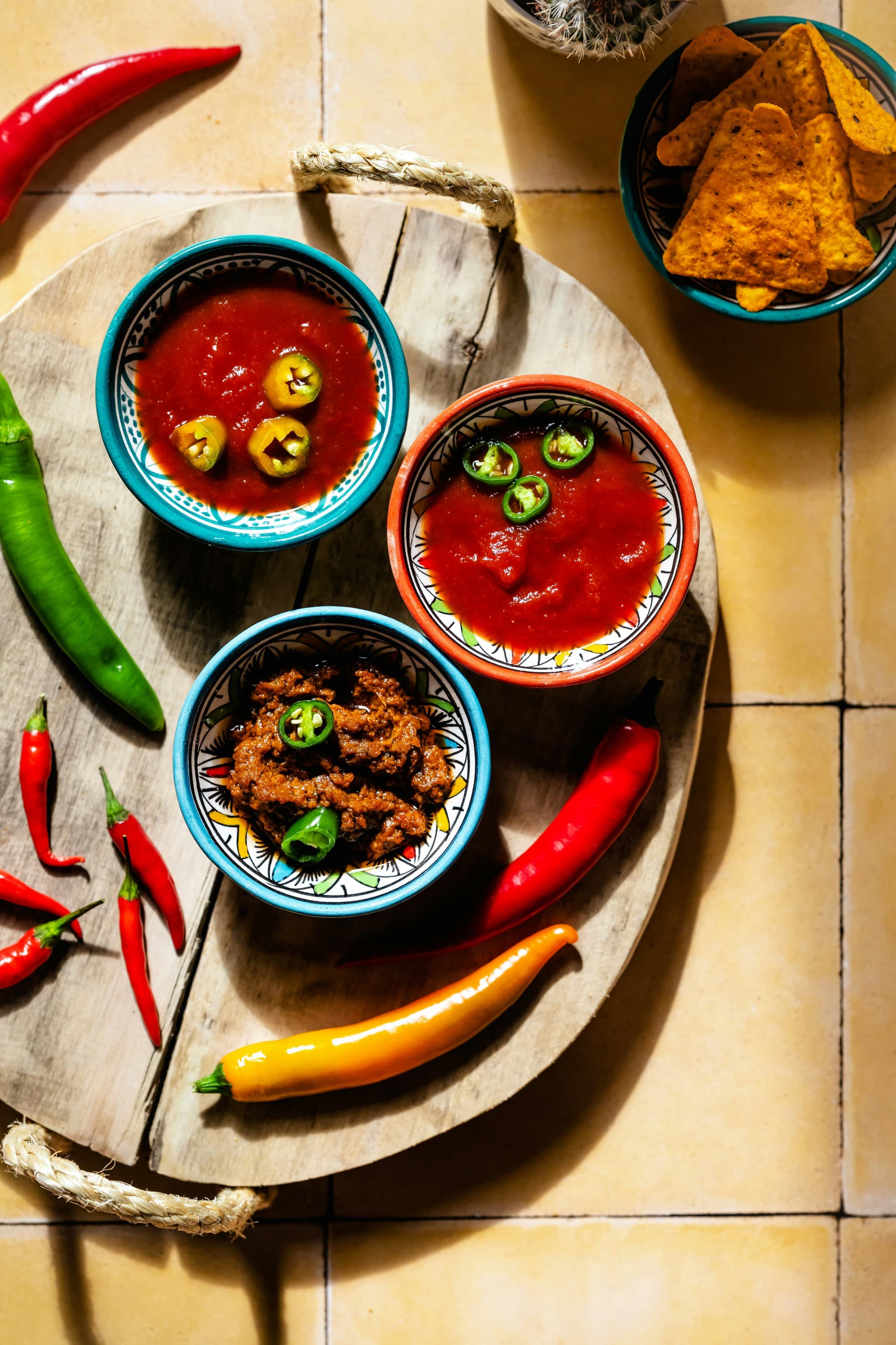 An aerial view of a wooden board with salsa bowls, chili peppers, and tortilla chips.