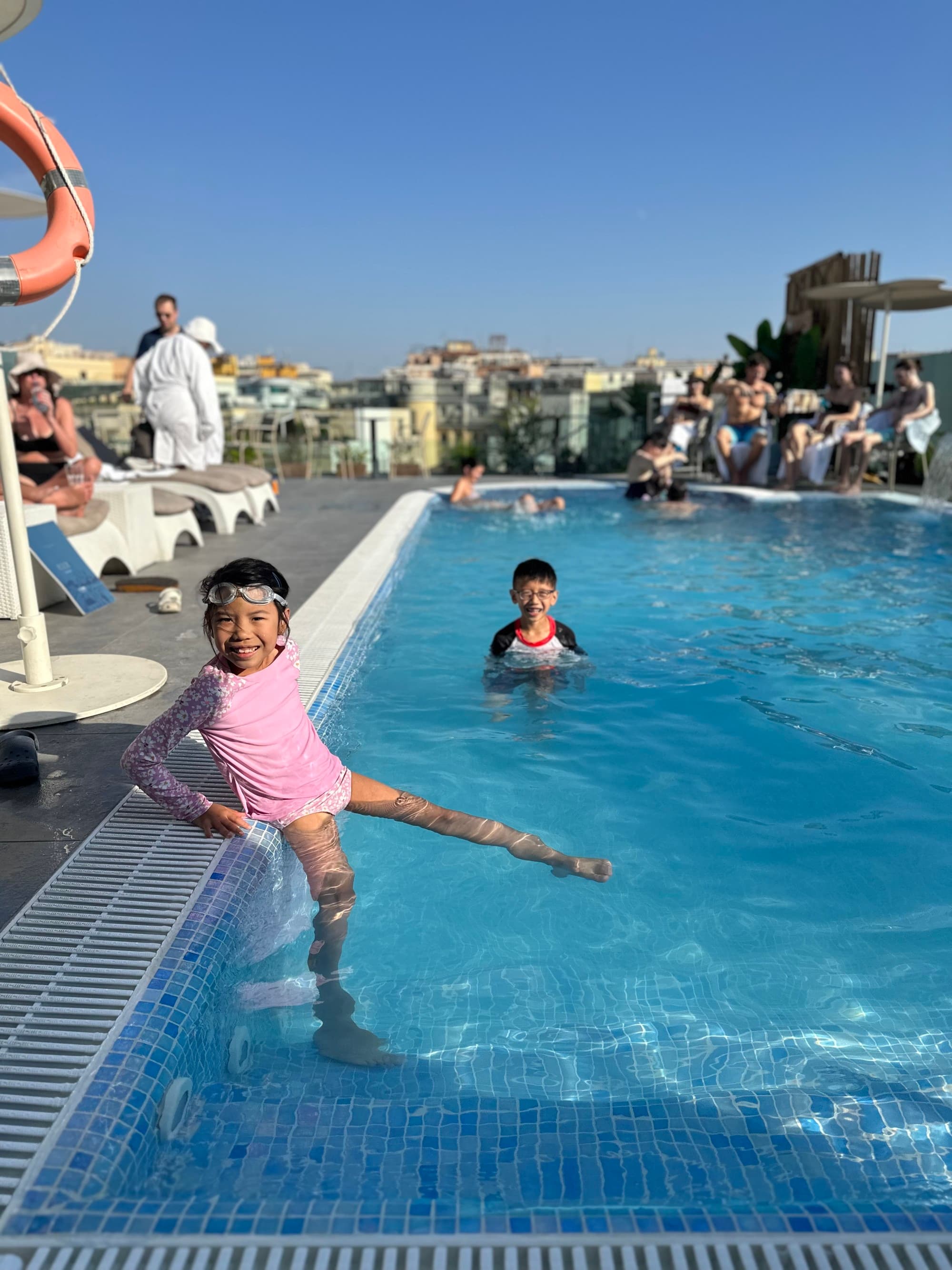 Two kids in a rooftop pool at one of the best hotels in Rome for families, enjoying a sunny day with the city skyline visible behind them.