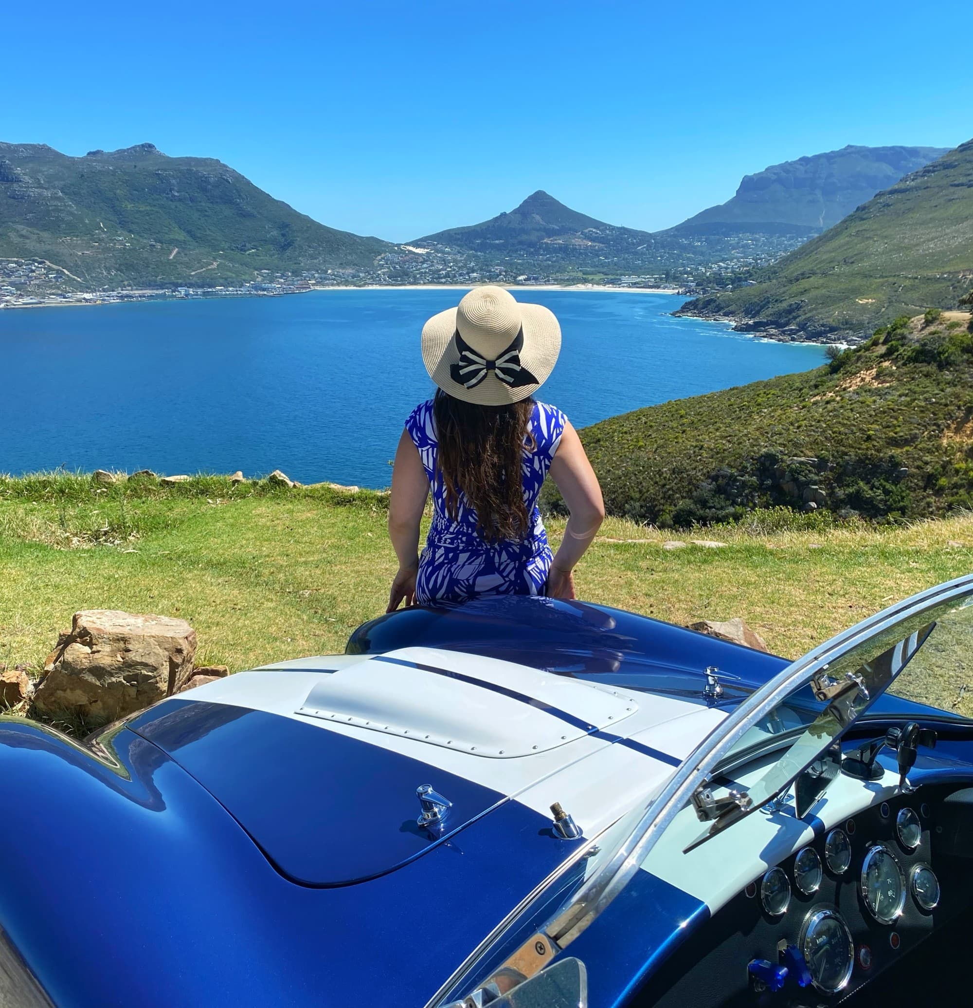 Courtney in a blue dress and sun hat sitting on the edge of a blue Cobra sports car overlooking a lake with mountains in the distance.