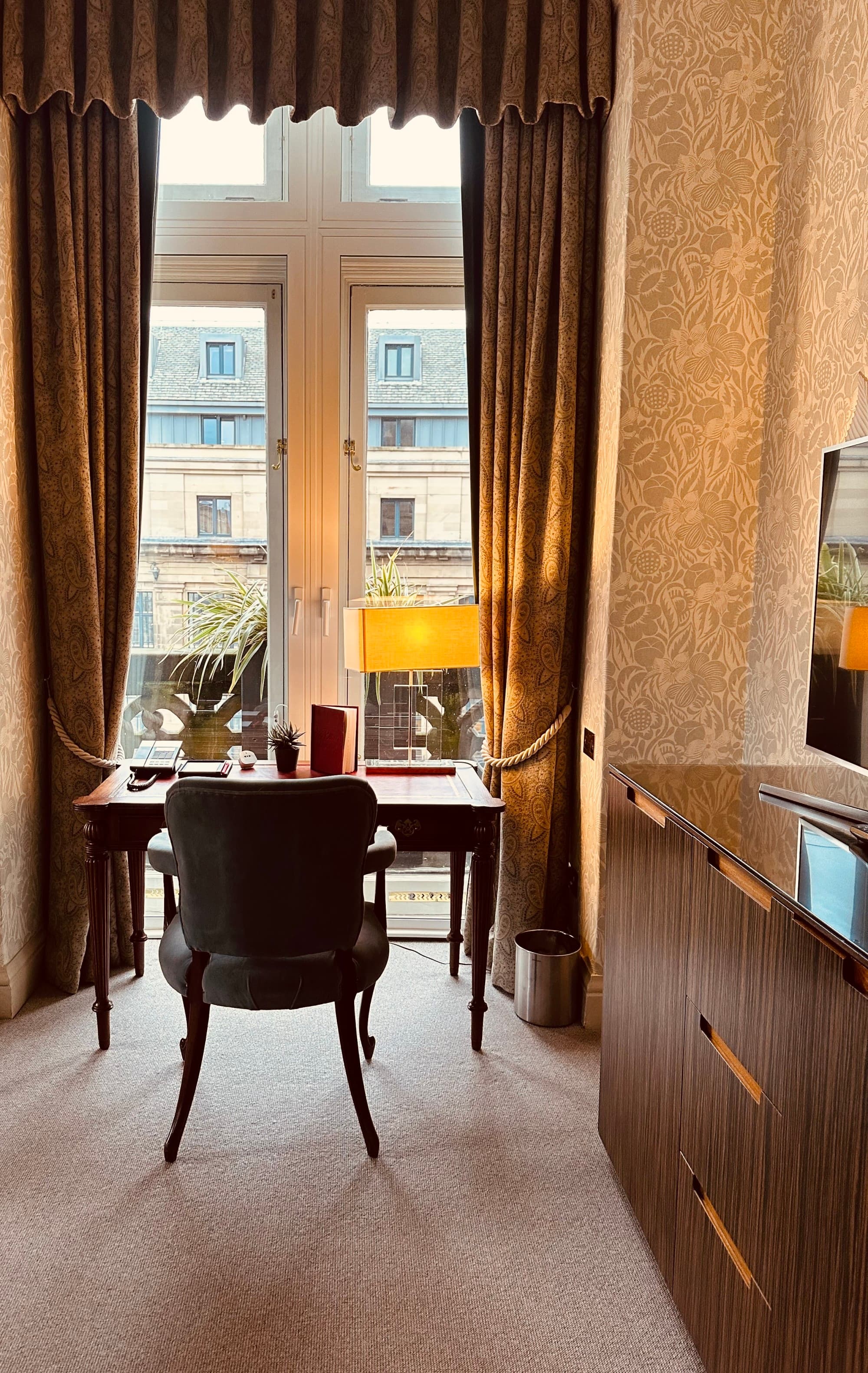 A television and desk / study area of a hotel room, facing a floor-ceiling window with brown curtains.