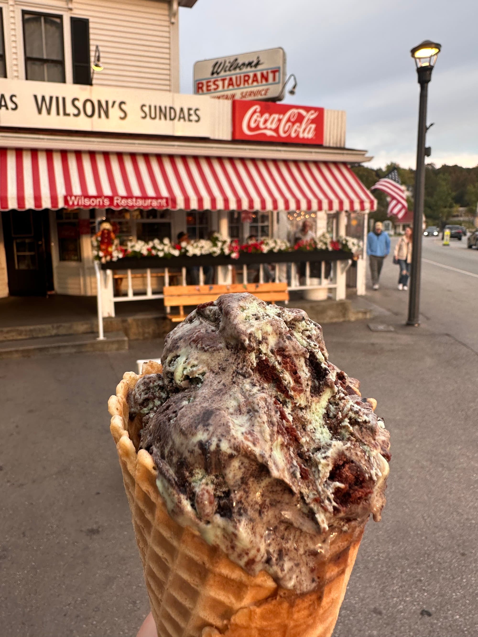 A close-up view of an ice-cream cone in front of a store with red and white awning.