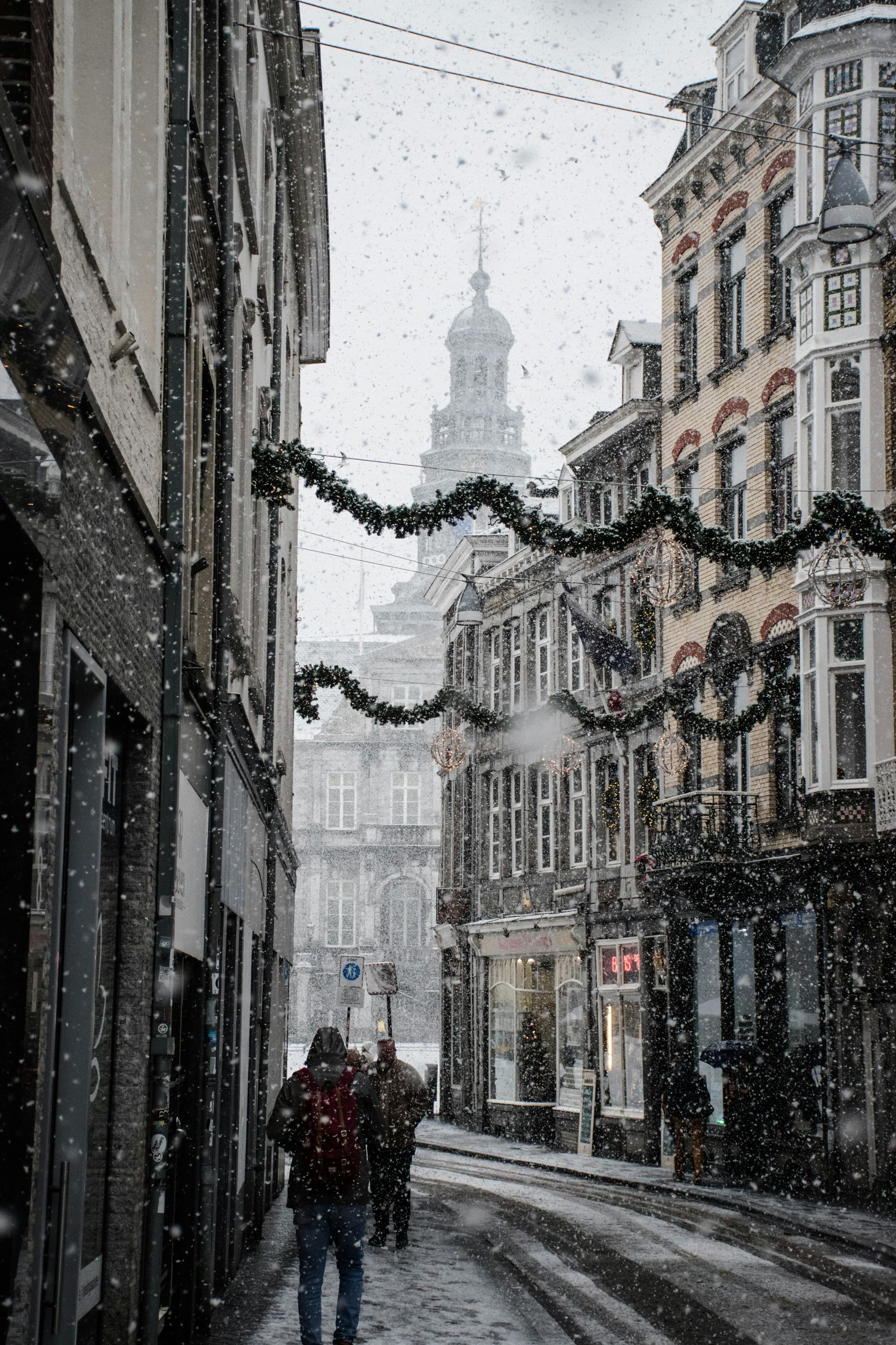 View of a narrow street with few pedestrians in light snow decorated with Christmas garlands above