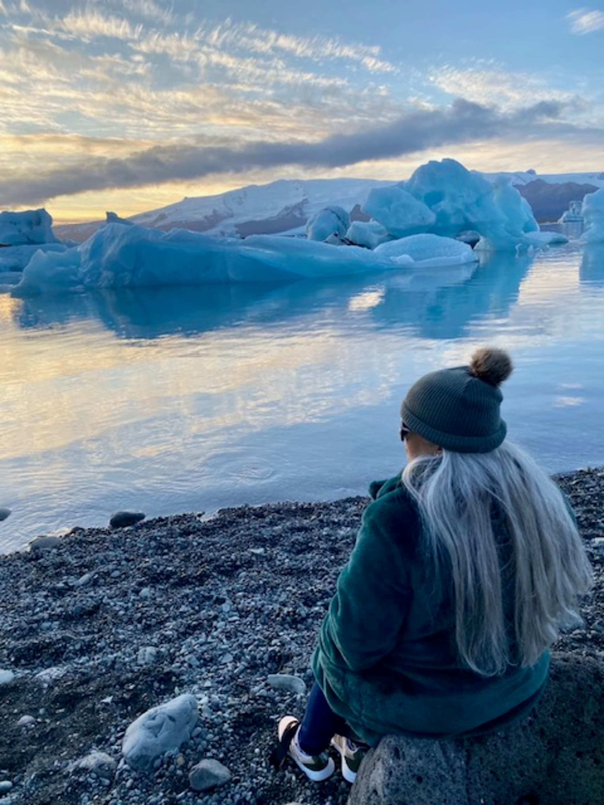 This image depicts a blonde woman sitting on a rocky shore and looking out onto a clear lake with ice bergs in the surrounding areas.