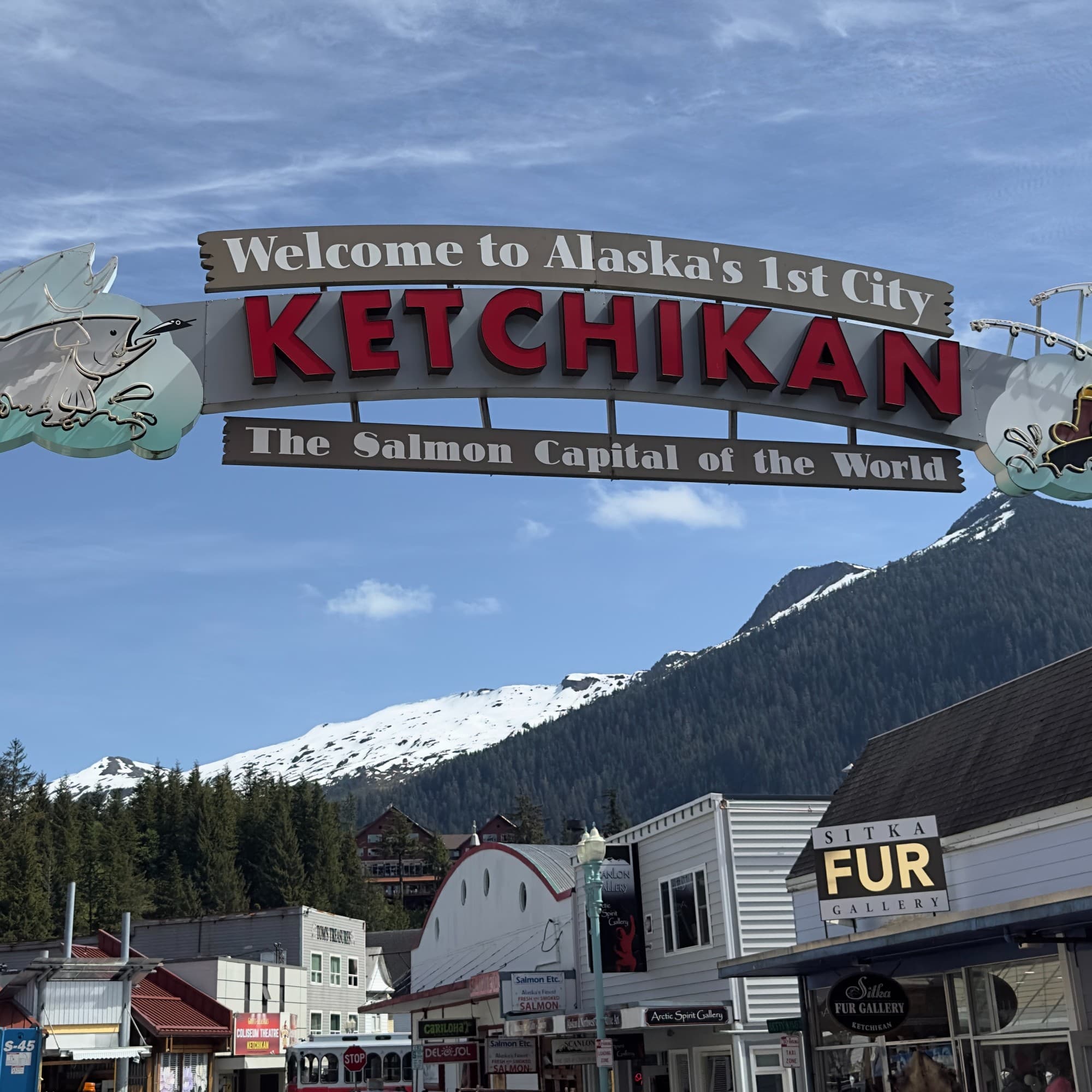 A welcome sign at the entrance to a town reading, " Welcome to Alaska's 1st City. Ketchikan. The Salmon Capital of the World." with snow-capped mountains in the distance.