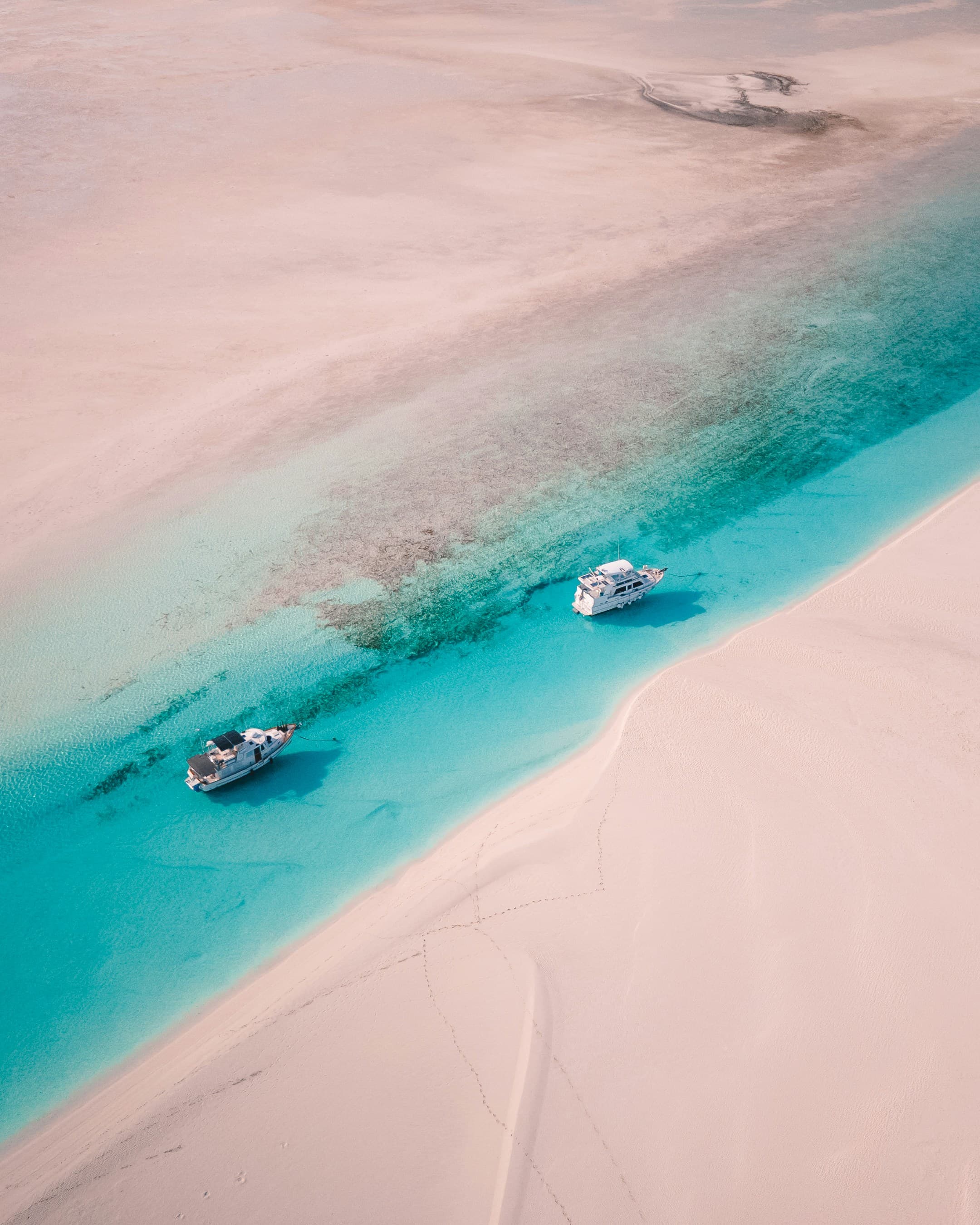 This image depicts an aerial view of two white boats on a body of water surrounded by white sand.