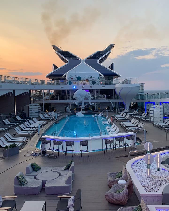 This image depicts a pool deck on a cruise ship at sunset. There are lounge chairs and an orange tinted sky in view.