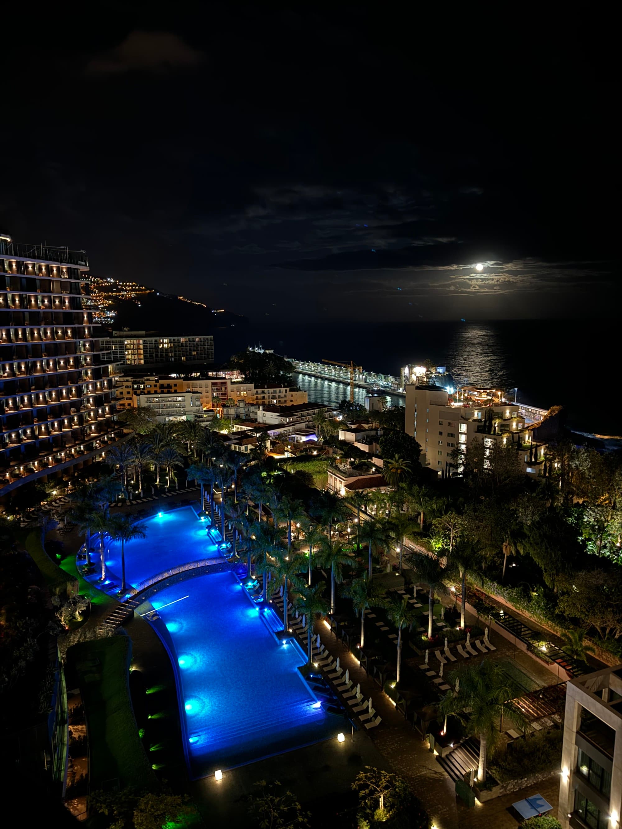 A view of Funchal City from the Savoy Palace hotel's balcony, overlooking the hotel property, coastline and ocean at night, with a full moon's light bouncing off of the water's surface.